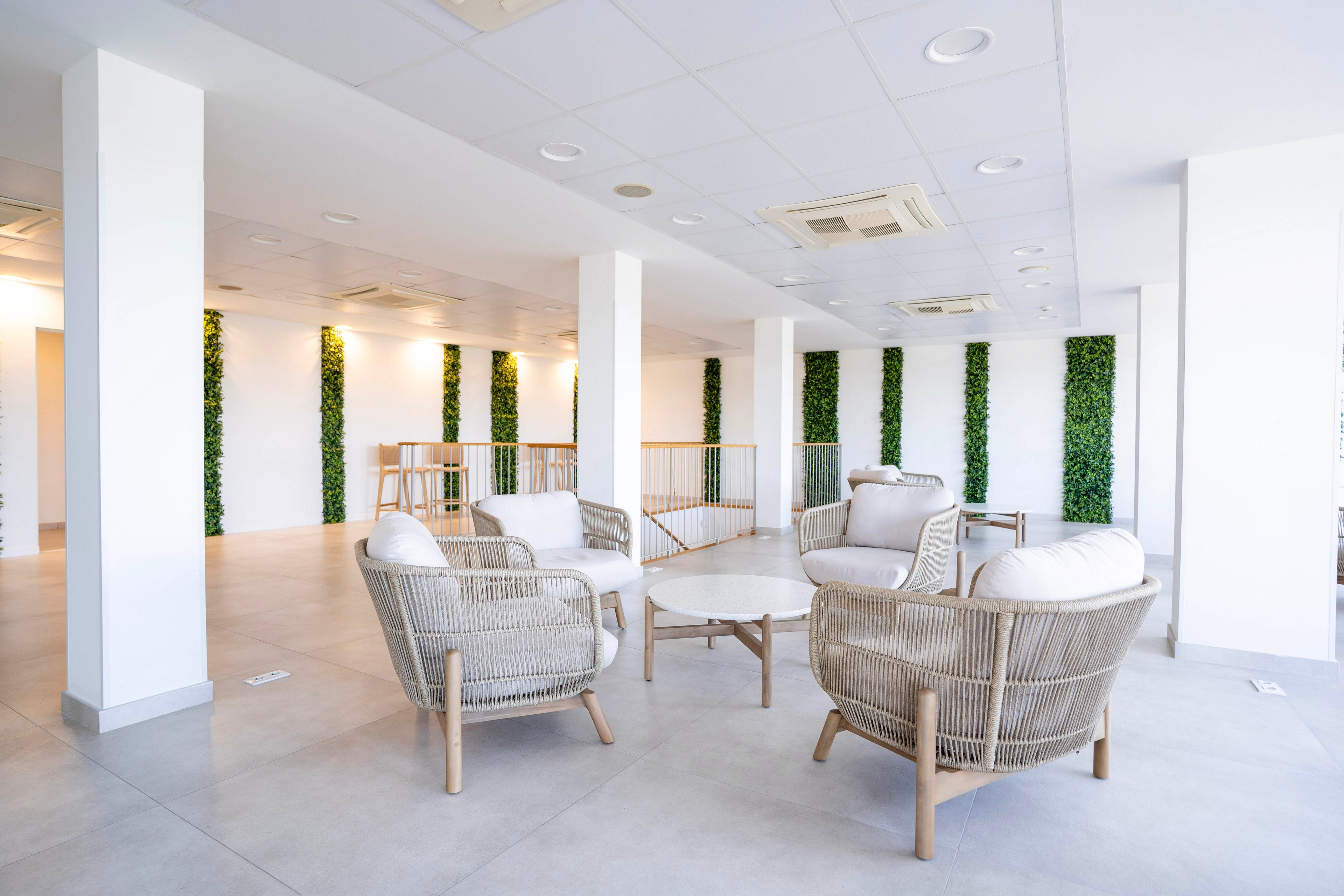 A bright modern lounge features a cluster of woven armchairs around a small circular table. The space is open-plan with white walls, light flooring, and a suspended ceiling with recessed lighting and air conditioning units. Vertical green plant panels add natural accents along the walls. In the background, a high table with stools suggests a casual meeting or coworking area.