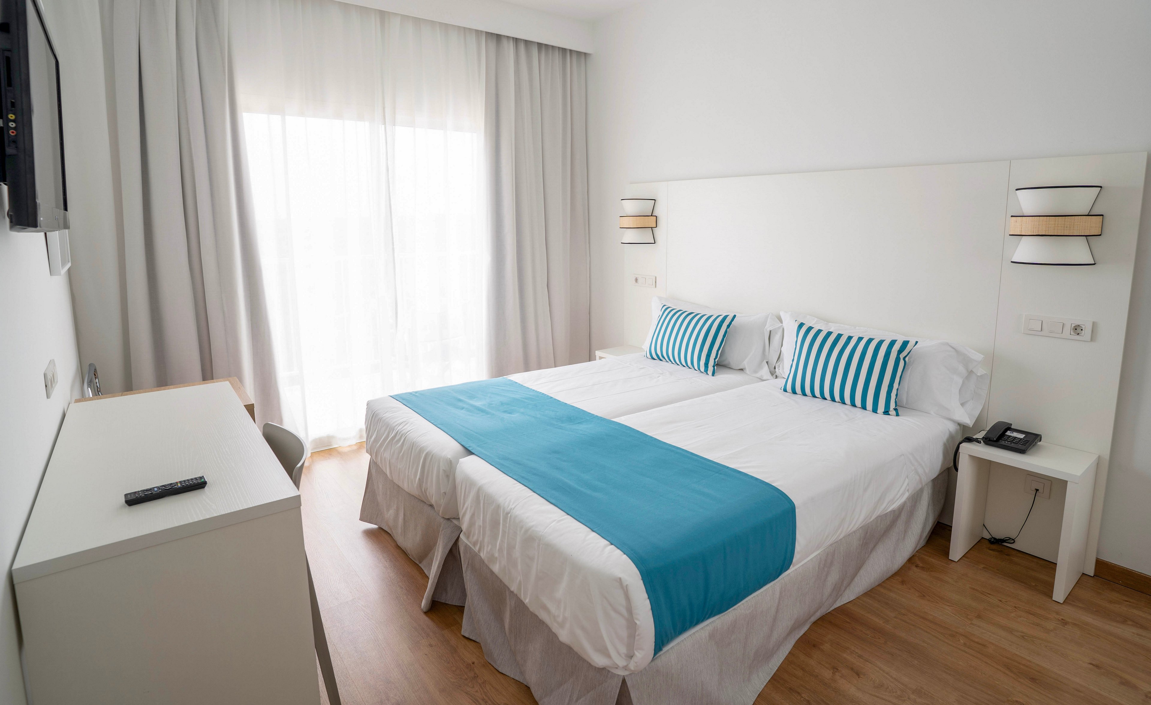 A bright, modern hotel room features two neatly made twin beds pushed together with white linens and turquoise runners. Striped turquoise and white pillows add a coastal accent to the minimalist decor. Sheer curtains cover a large window or balcony door, allowing soft natural light to fill the space. A bedside table with a telephone and a simple desk with a remote control complete the clean, contemporary look.