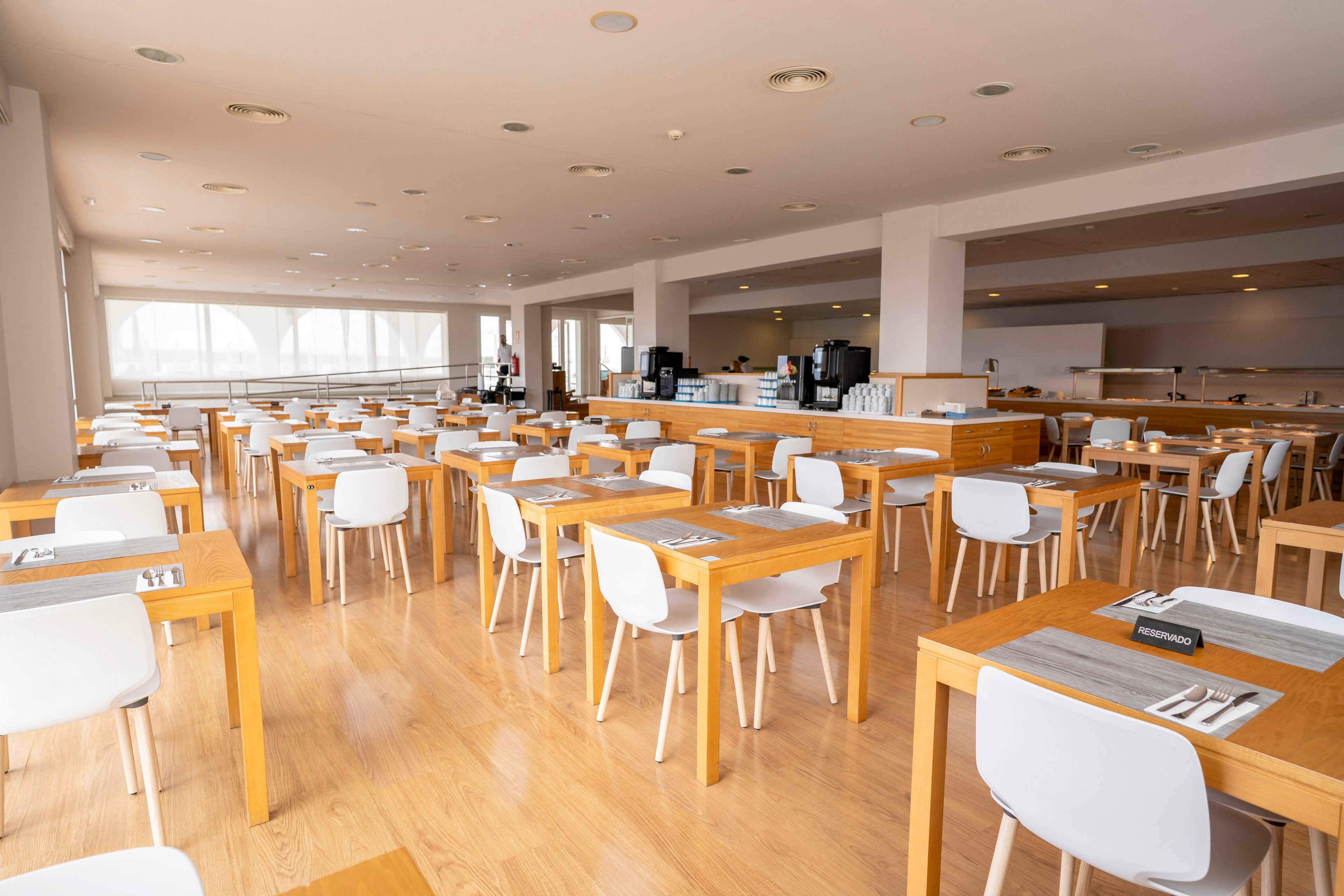 A spacious cafeteria is filled with rows of light wooden tables and white chairs. The room is empty of people, with neatly arranged place settings on each table. Large windows along one side let in abundant natural light, creating an airy atmosphere. The background shows a self-service counter and additional seating areas, emphasizing a clean and modern dining space.