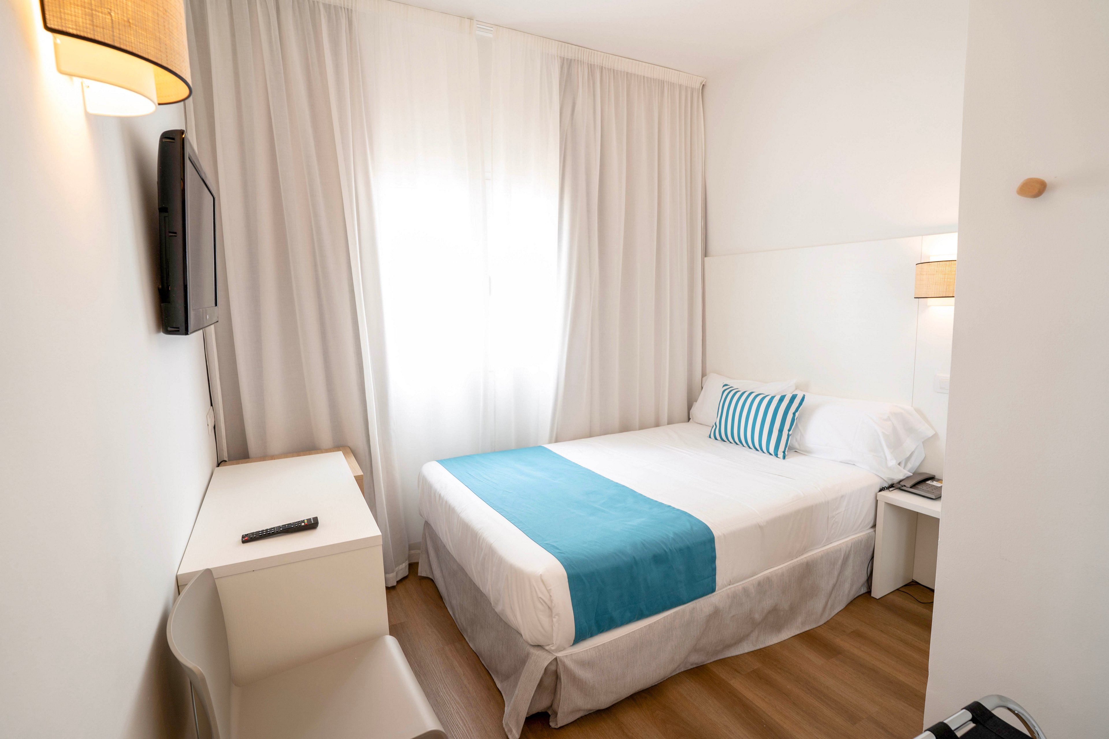 Compact modern hotel room featuring a neatly made single bed with white linens and a turquoise runner. The space is bright, with natural light filtering through sheer white curtains covering a large window. A wall-mounted flat-screen TV faces the bed, and a small desk with a chair sits beneath it. Warm wooden flooring and simple bedside tables with lamps create a clean, minimalist atmosphere.