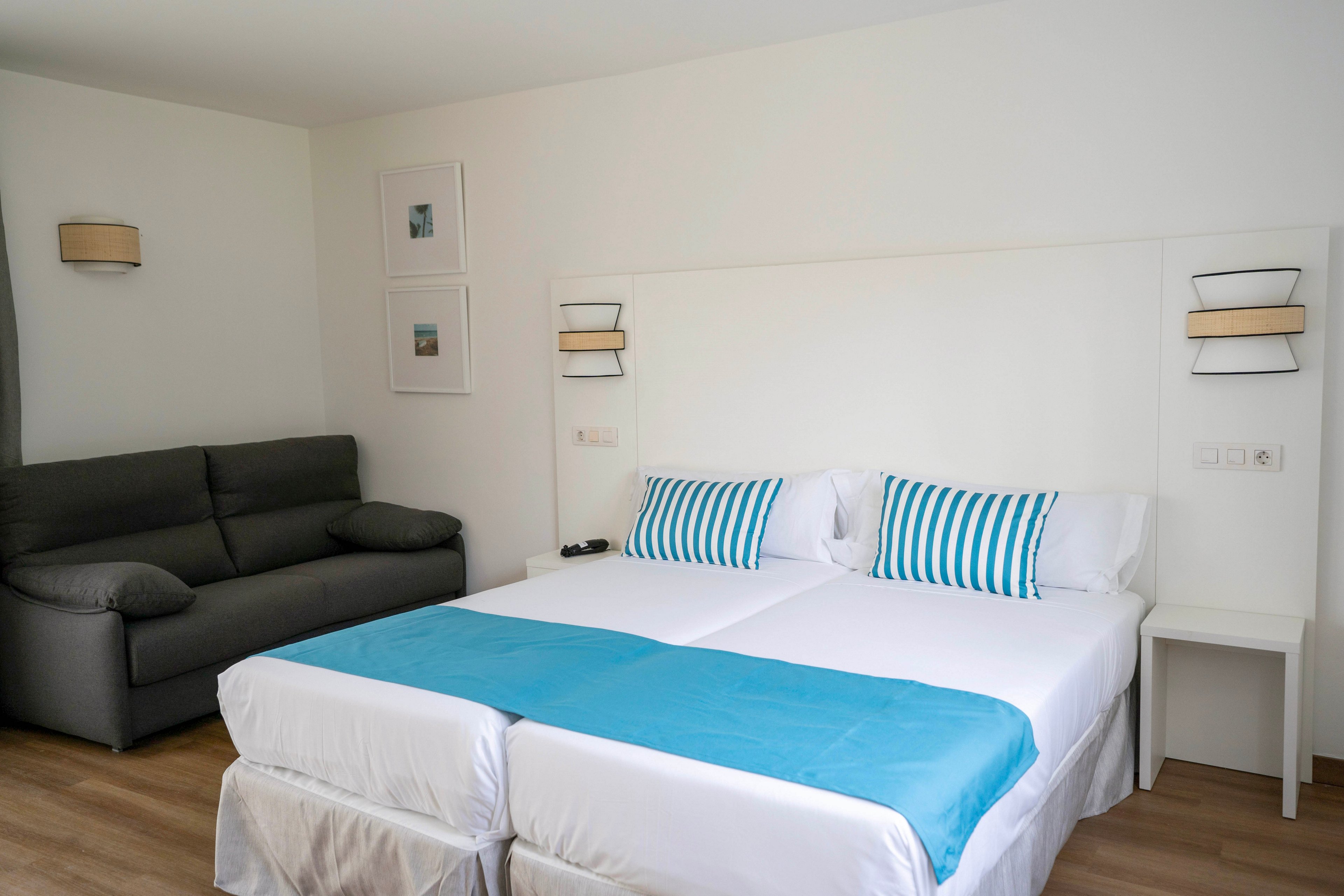 A bright, modern hotel room features two neatly made twin beds pushed together with white linens and turquoise bed runners. Matching striped turquoise and white pillows add a coastal accent to the minimalist decor. A dark gray sofa sits against the wall opposite the beds, providing additional seating. Light wood flooring, simple wall lamps, and framed artwork contribute to the clean and contemporary atmosphere.