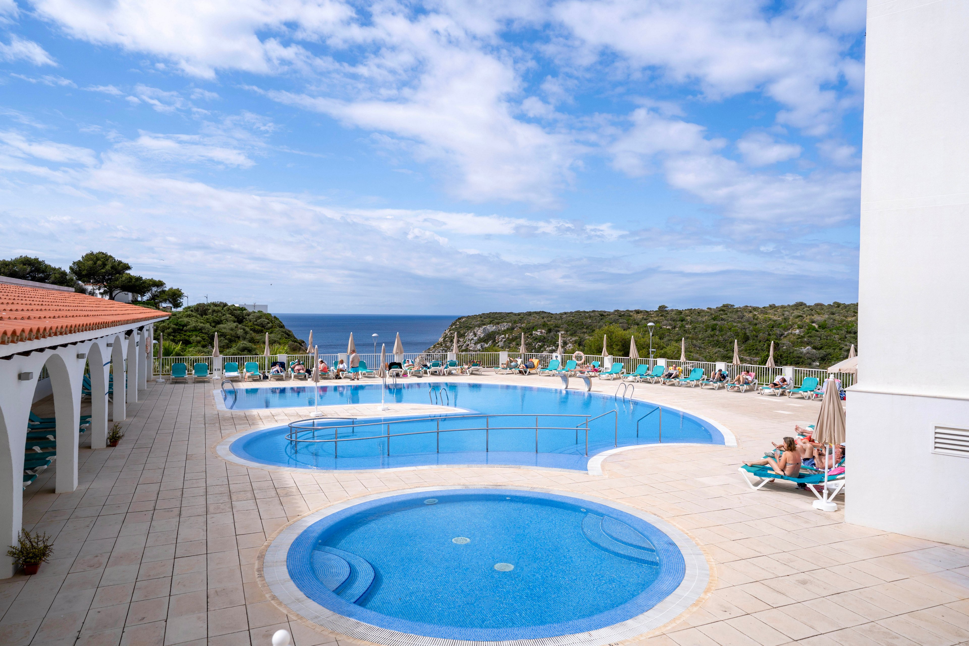 A large circular swimming pool and smaller kiddie pool sit on a spacious tiled terrace at a seaside resort. Numerous guests relax on sun loungers under umbrellas around the pool area. White arches and buildings frame the scene, leading the eye toward the blue sea and distant rocky coastline. The sky is partly cloudy, creating a bright yet relaxed holiday atmosphere.