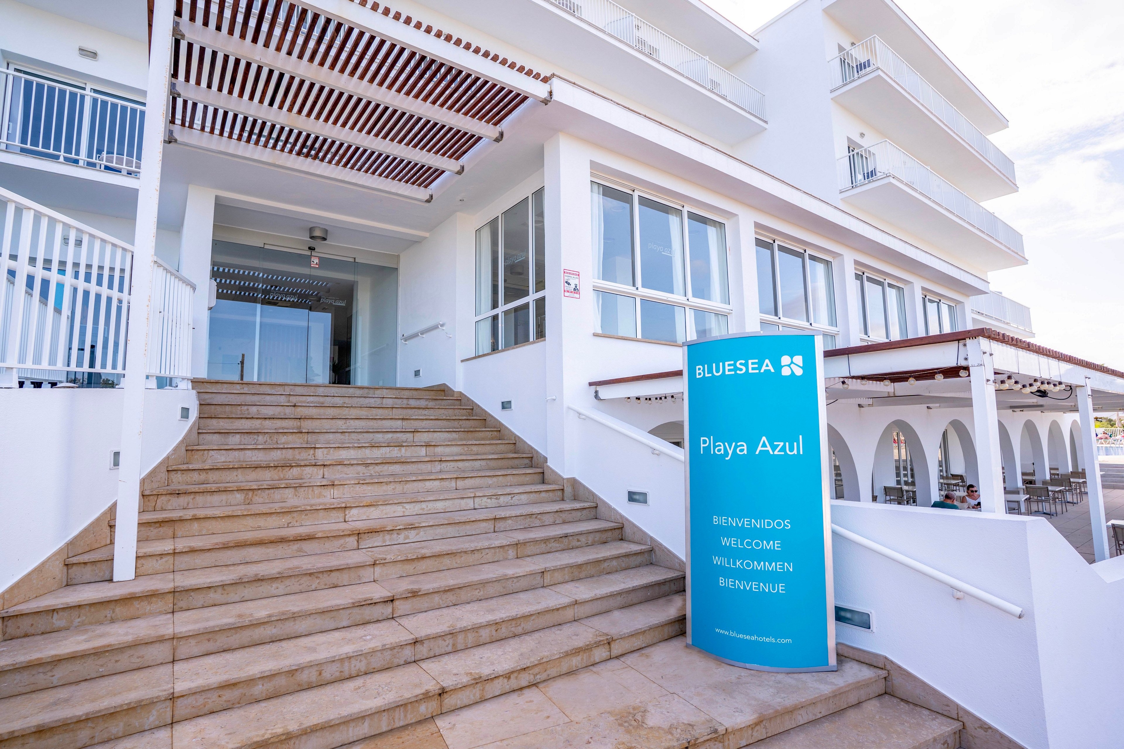 Exterior view of the Blue Sea Playa Azul hotel entrance with wide stone steps leading up to glass doors. A tall blue sign stands beside the staircase displaying the hotel name and multilingual welcome text. The white building features balconies, railings, and shaded terraces under a clear sky. The scene suggests a coastal or resort setting, emphasizing hospitality and vacation travel.