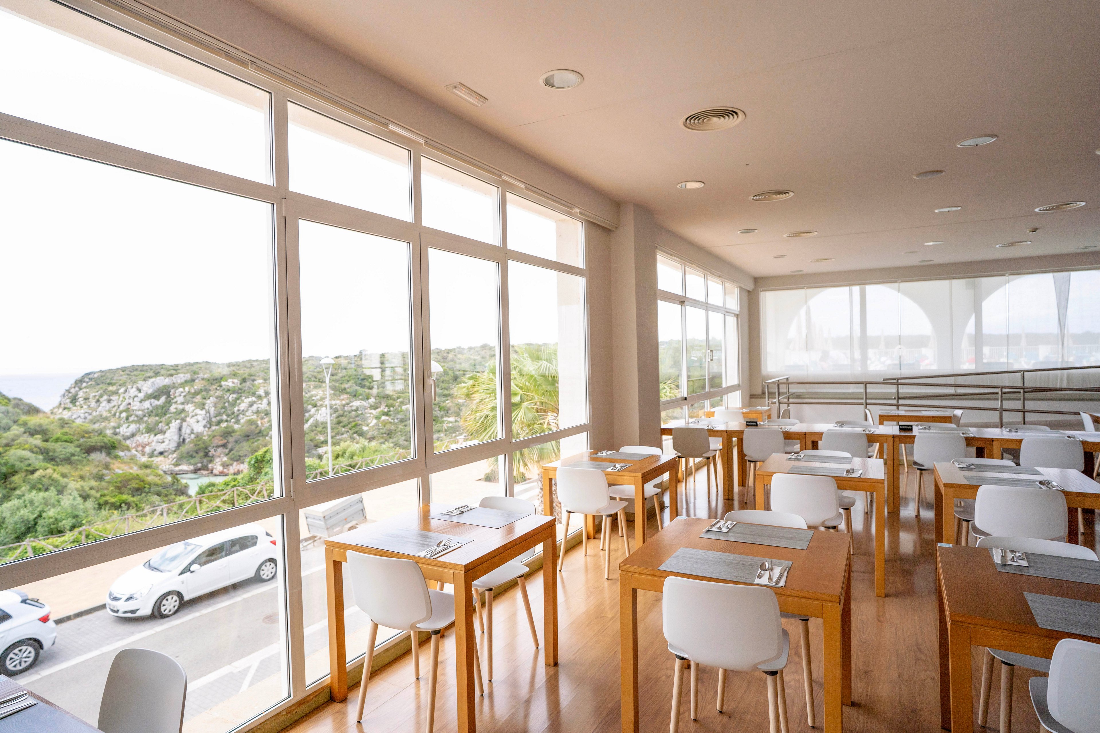 Spacious modern restaurant interior with wooden tables and white chairs is shown in daylight. Large floor-to-ceiling windows reveal a scenic coastal landscape with greenery, cliffs, and parked cars outside. Place settings with placemats and cutlery are neatly arranged on each table, suggesting readiness for guests. The atmosphere feels airy, clean, and relaxed, ideal for casual dining with a view.