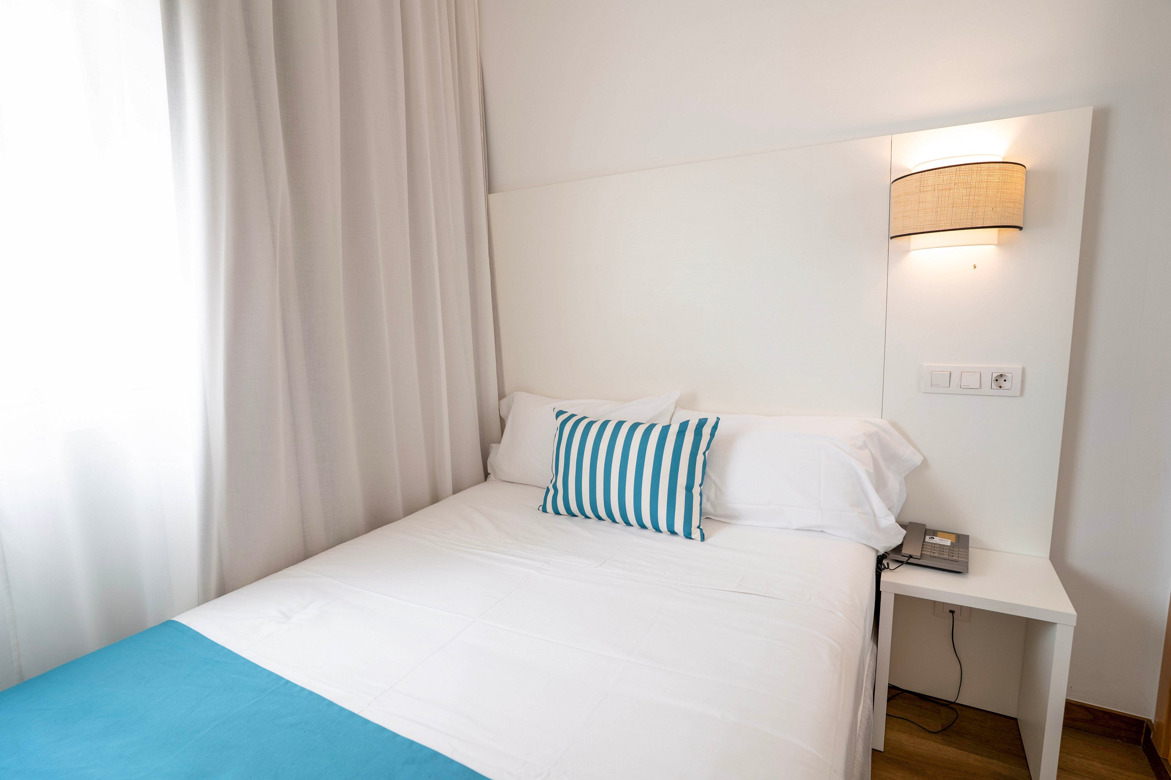 A small, neatly made bed sits against a white wall in a minimalist bedroom. The bed features crisp white linens accented by a turquoise bed runner and a blue-and-white striped pillow. Soft daylight filters through long white curtains on the left side of the room. A wall-mounted lamp and a compact bedside table with a phone complete the simple, modern decor.