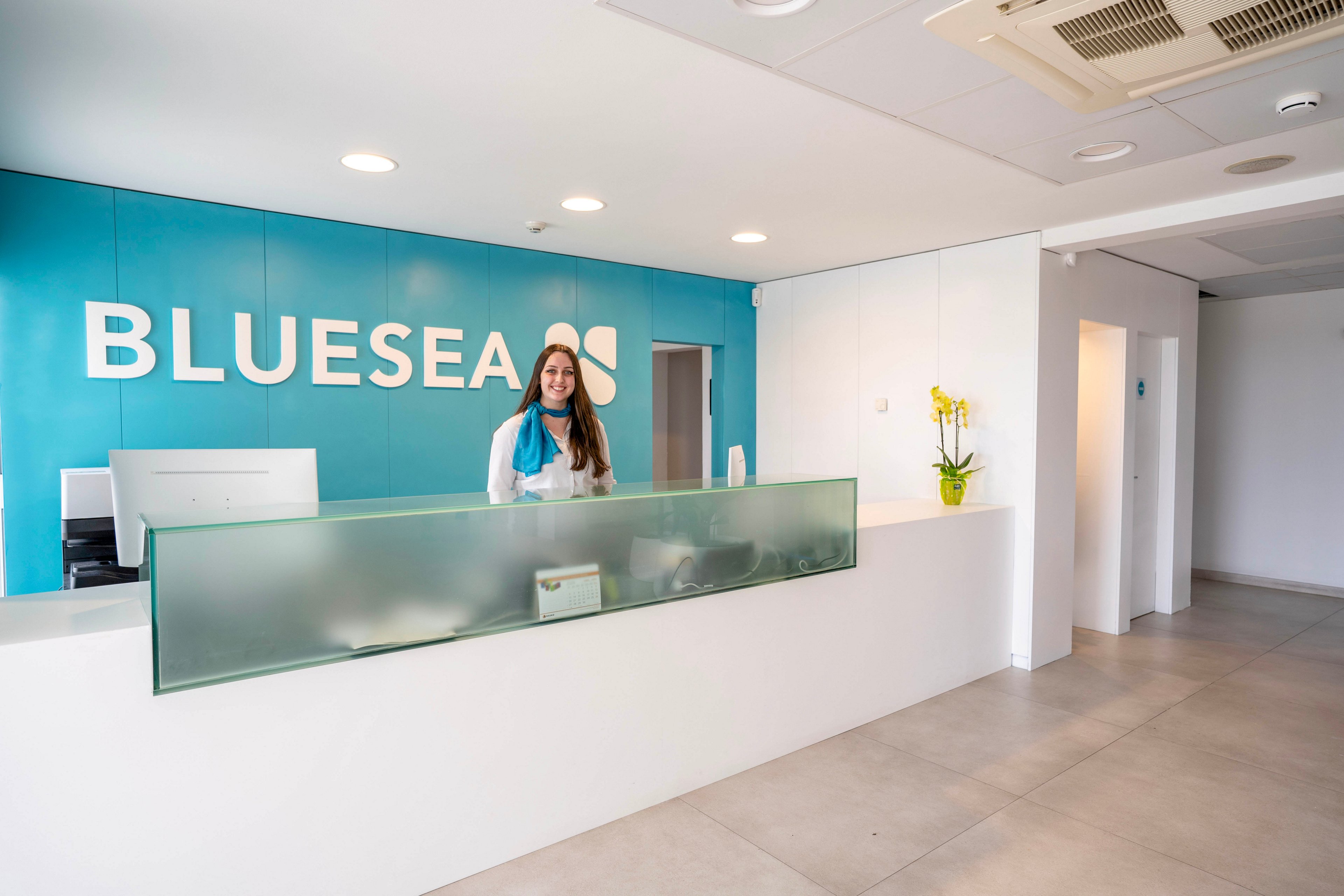 A smiling female receptionist stands behind a sleek white and glass front desk in a modern reception area. The turquoise back wall prominently displays the word BLUESEA with a simple graphic mark. The space is bright and minimal, with neutral flooring, white walls, and a ceiling-mounted air conditioning unit. A small vase with yellow flowers decorates the counter, and a hallway with doors is visible in the background.