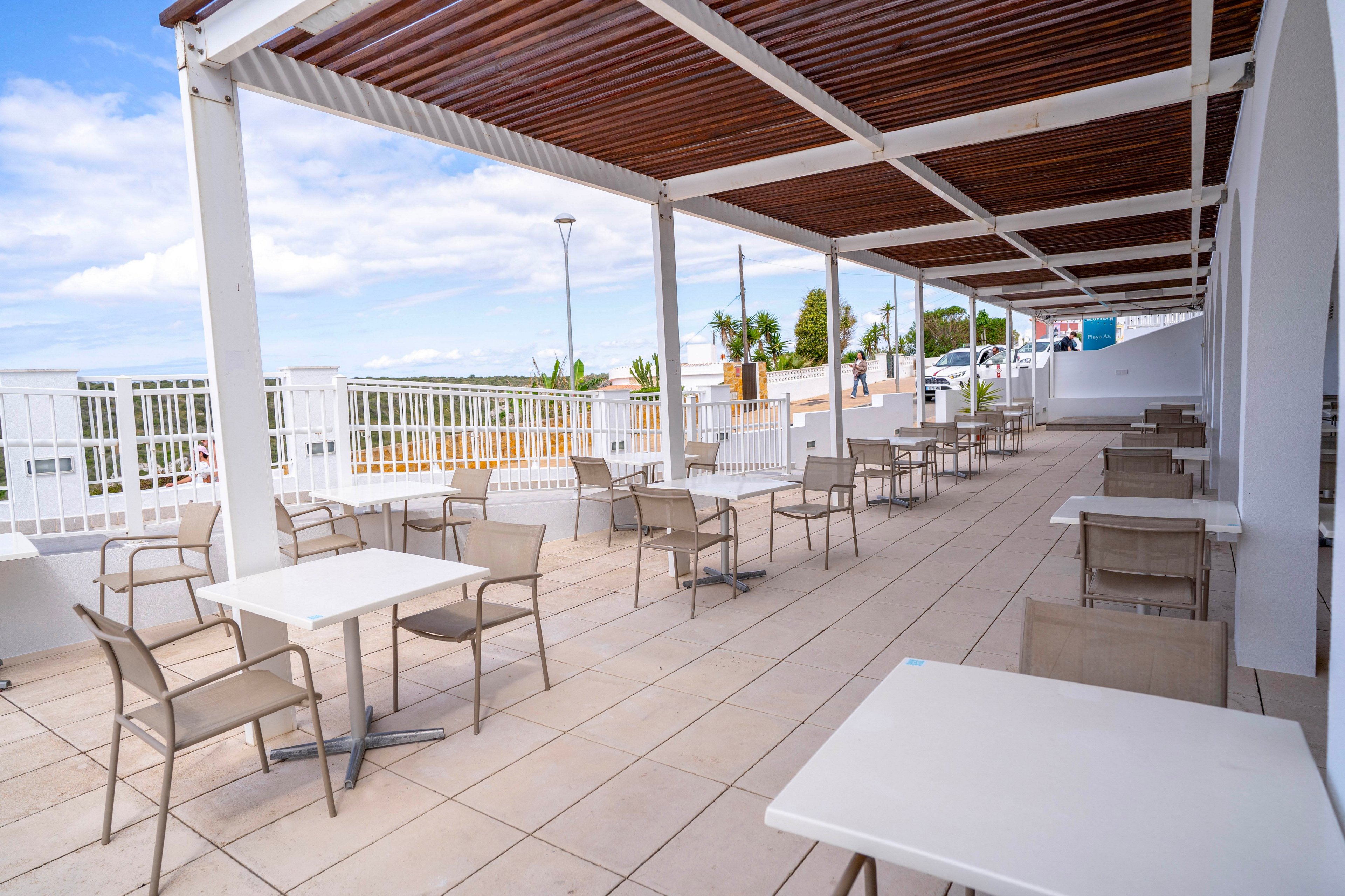 An empty outdoor terrace cafe features rows of tables and chairs under a modern pergola. The space is tiled in light stone and bordered by white railings overlooking a bright, open area. A few streetlights and distant greenery are visible beyond the terrace, suggesting a warm, pleasant day. The scene feels clean, minimal, and ready for guests.