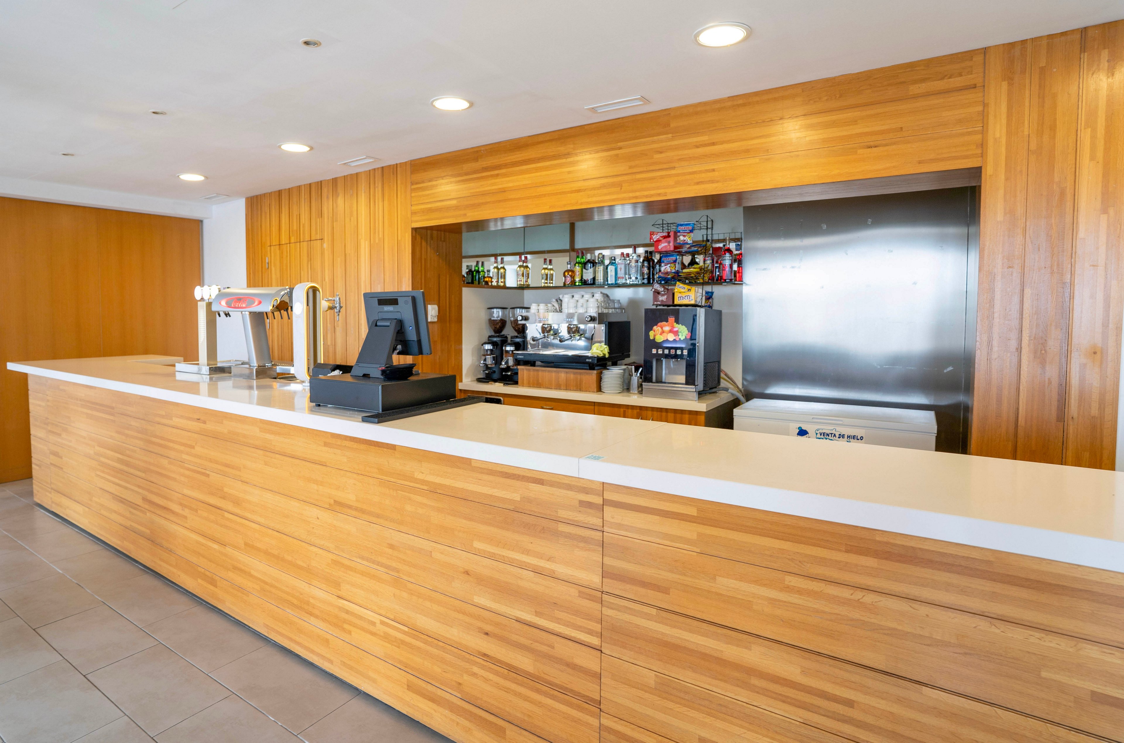 A modern bar counter with a long white surface and light wooden paneling dominates the scene. Behind the counter, shelves display various bottles and glasses along with a coffee machine and beverage dispensers. The setting appears clean and minimalistic, with stainless steel equipment and neutral tiled flooring. No customers or staff are present, emphasizing the tidy, contemporary design of the bar area.