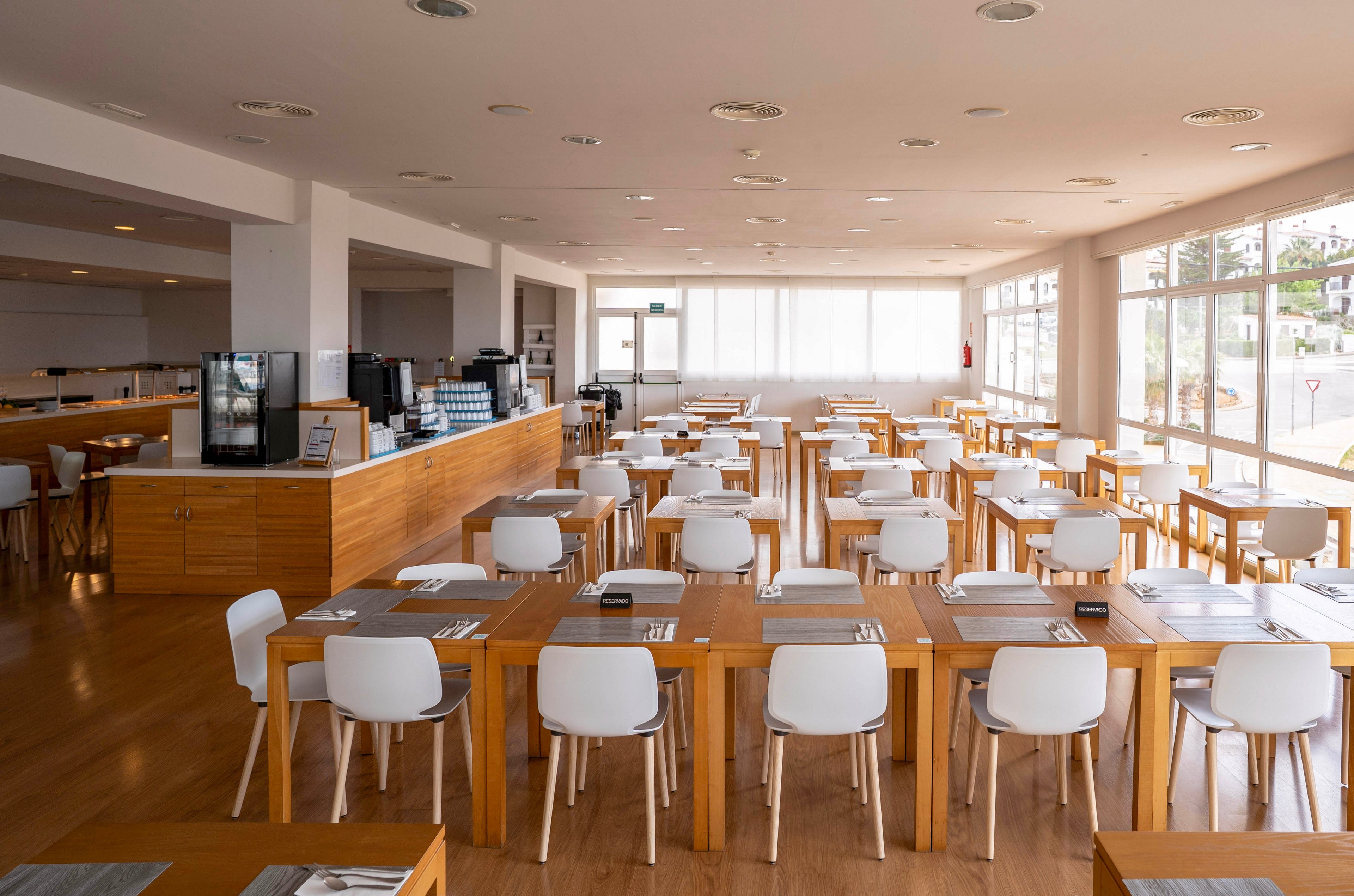 A spacious, modern cafeteria features rows of wooden tables neatly arranged with white chairs. Place settings with cutlery and placemats are prepared on each table, suggesting readiness for guests. Large windows on two sides fill the room with natural light, creating an airy and inviting atmosphere. A long service counter with beverage machines and equipment runs along one side of the space.