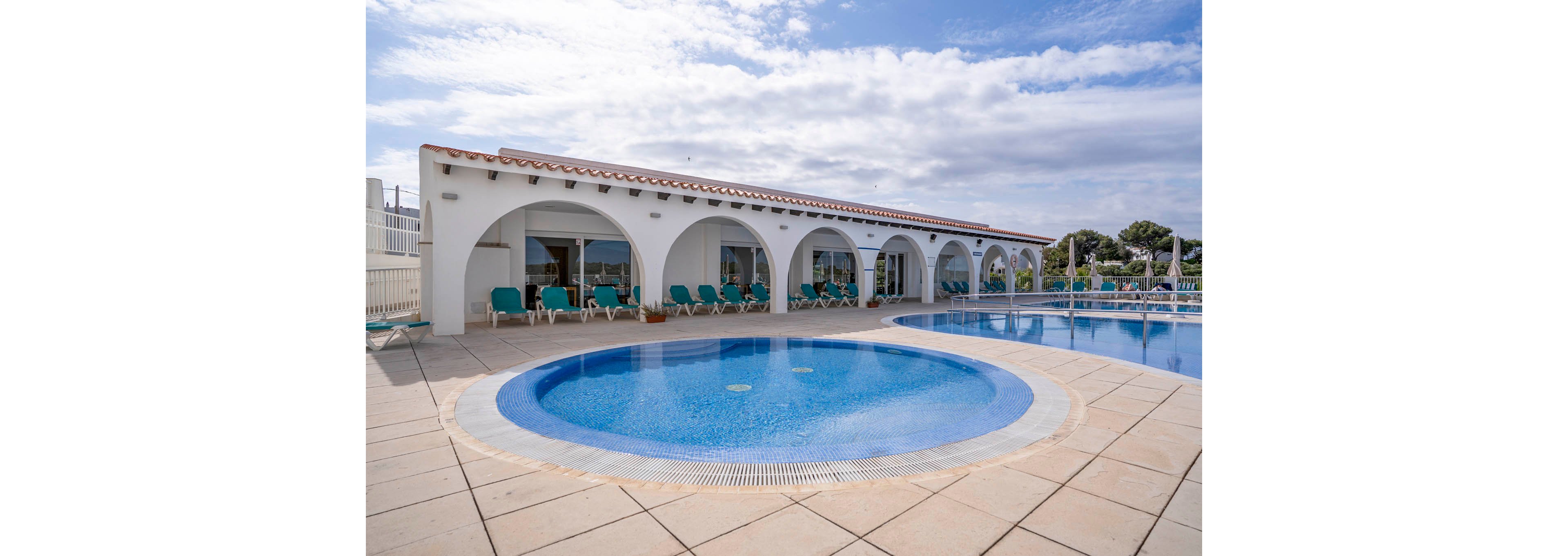 A circular shallow pool sits in the foreground of a sunny resort terrace. Behind it, a long white building with multiple arches opens onto a row of turquoise lounge chairs. A larger swimming pool extends along the right side, reflecting the blue sky and scattered clouds. The scene suggests a relaxed holiday atmosphere at a coastal or Mediterranean-style hotel.