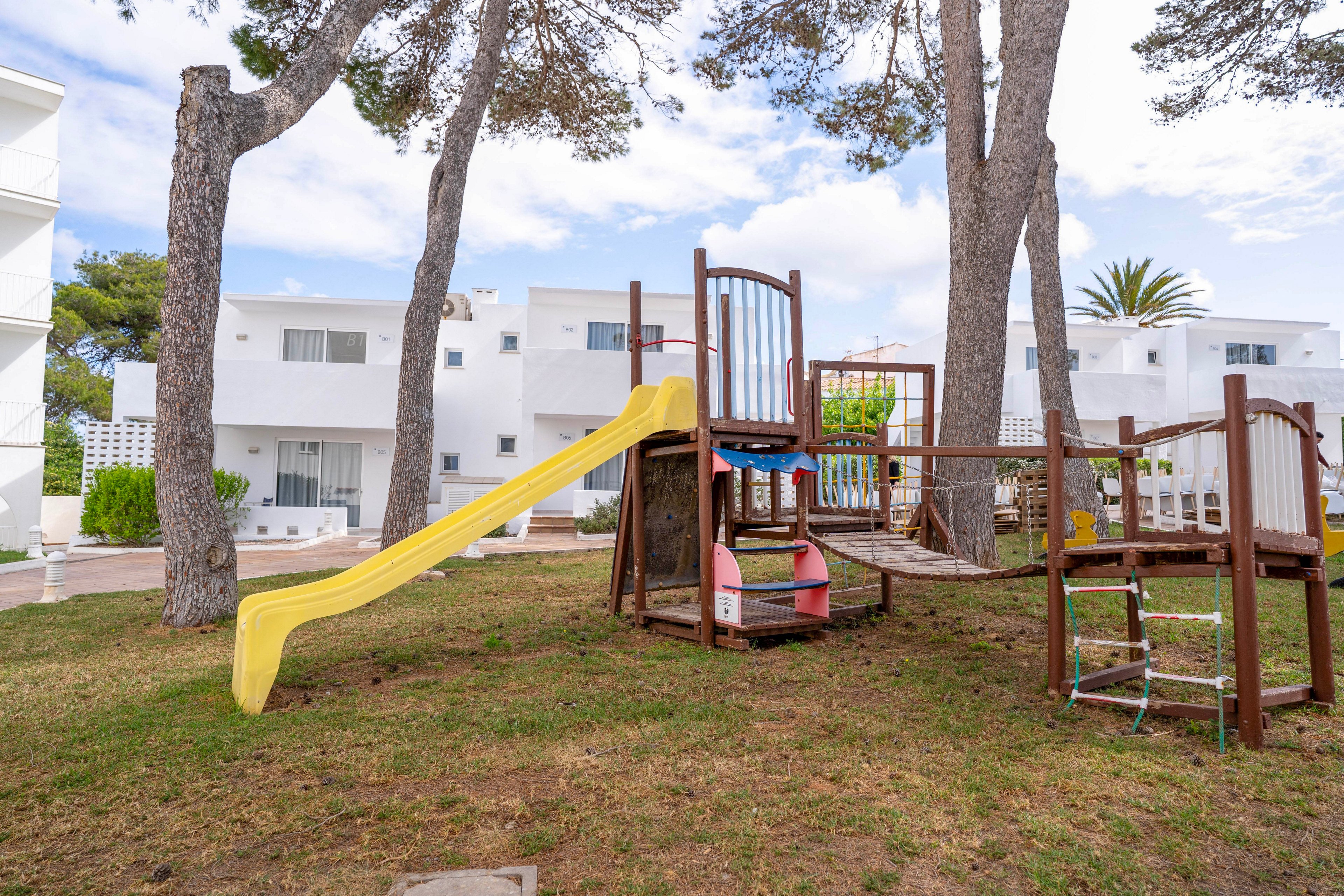 A small wooden playground structure with a yellow slide stands on a grassy courtyard. The play area includes a climbing ladder, platform, and small playhouse elements beneath the structure. Tall trees surround the playground, providing shade and framing the scene. In the background, white modern residential buildings with balconies and windows create a calm neighborhood setting under a partly cloudy sky.