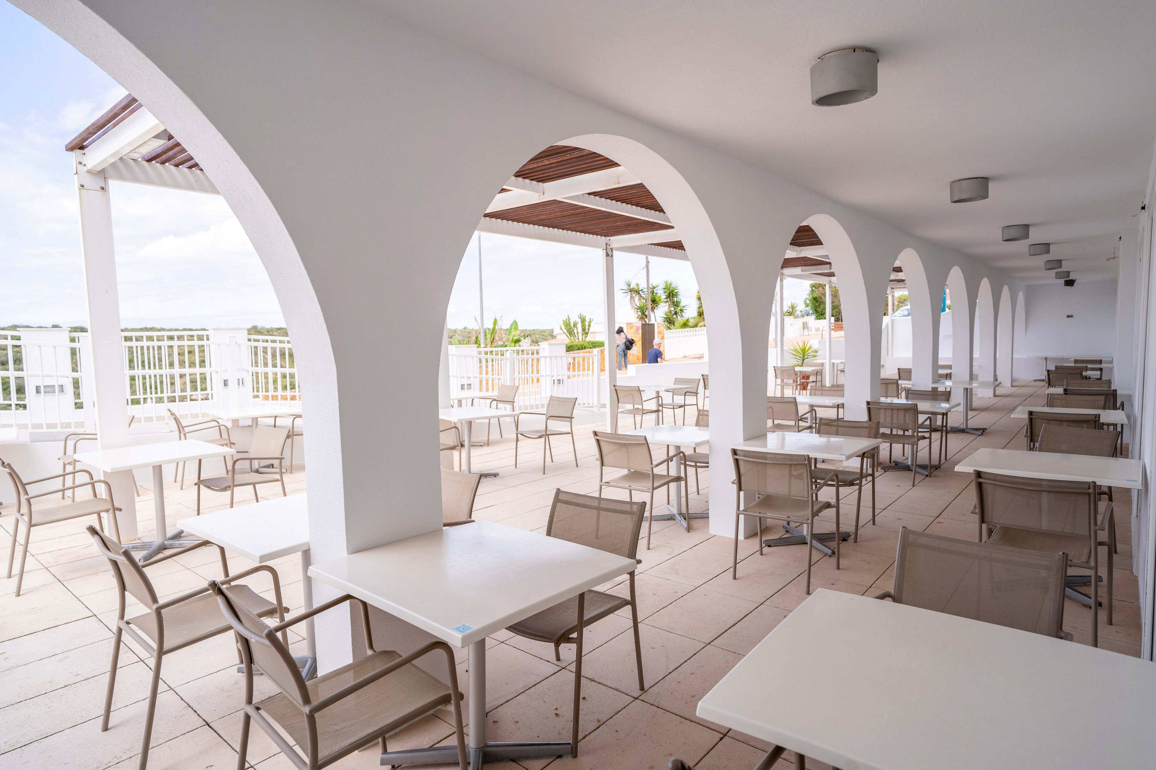 An open-air terrace café features rows of white arches and modern tables with beige chairs. The space is bright and airy, with a tiled floor and a partially covered ceiling. In the background, greenery and a distant figure add depth to the scene. The overall atmosphere feels relaxed and inviting, suitable for outdoor dining or social gatherings.