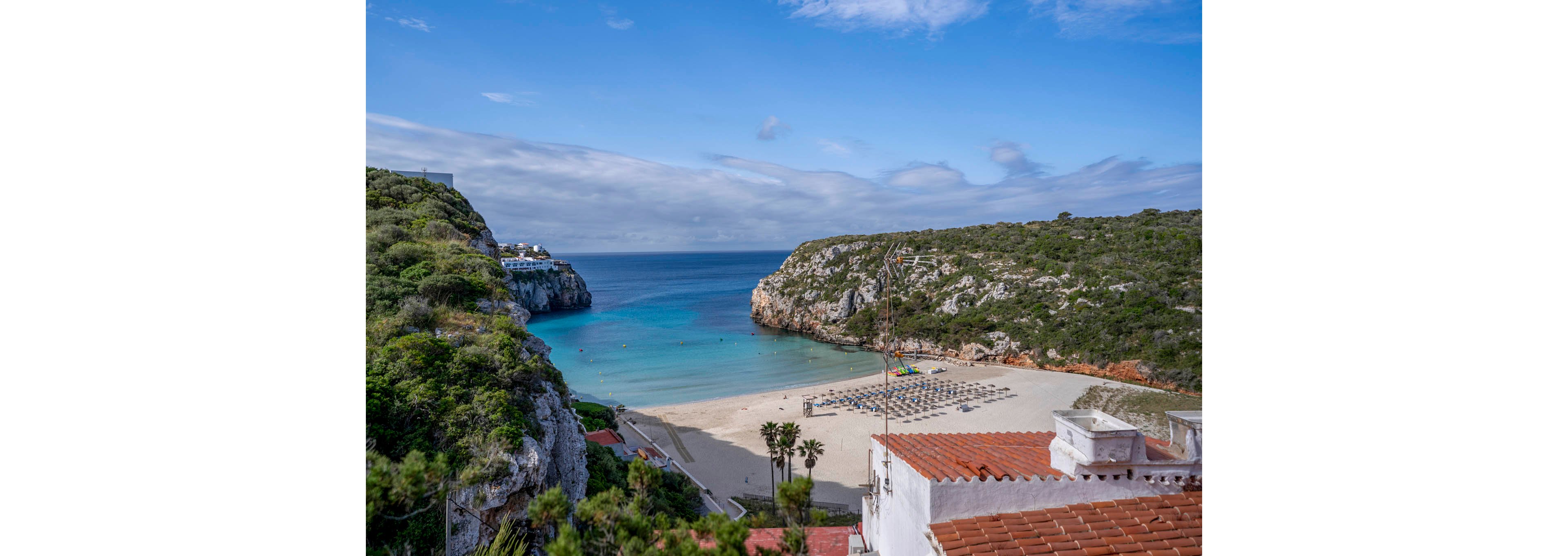 A secluded sandy beach sits between two rocky, green-covered cliffs along a calm turquoise sea. Neatly arranged rows of empty sun loungers and umbrellas line the shoreline, suggesting a quiet resort setting. Whitewashed buildings with terracotta roofs overlook the bay from the foreground. Soft clouds stretch across the blue sky, adding to the tranquil coastal atmosphere.