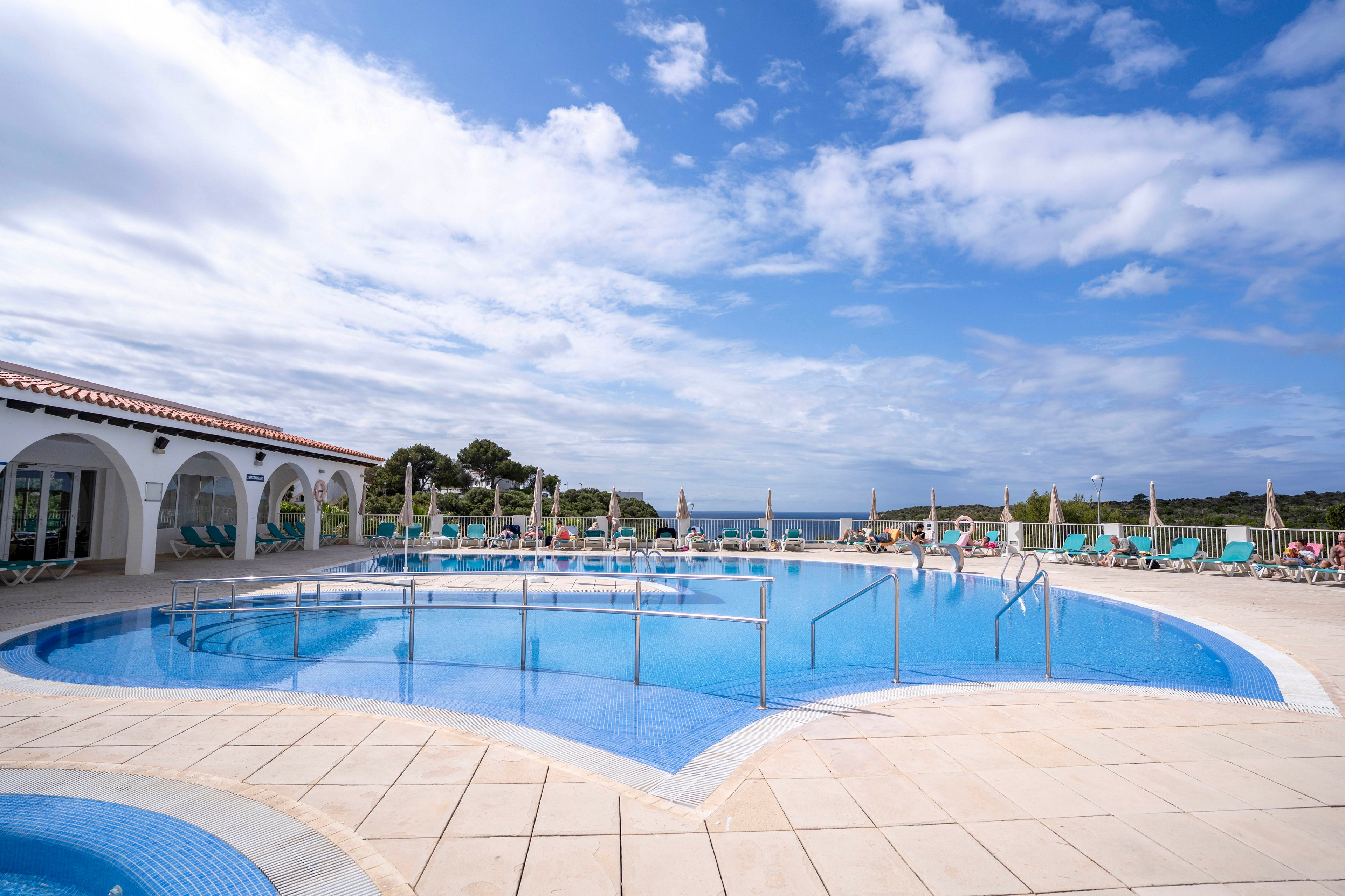 A large resort swimming pool sits at the center of an open-air terrace. White tiled decking surrounds the pool, with sun loungers and umbrellas arranged neatly around the edges. A low white building with arches lines one side, while the sea and distant shoreline are visible beyond the pool area. The bright blue sky with scattered clouds enhances the relaxed, vacation atmosphere.