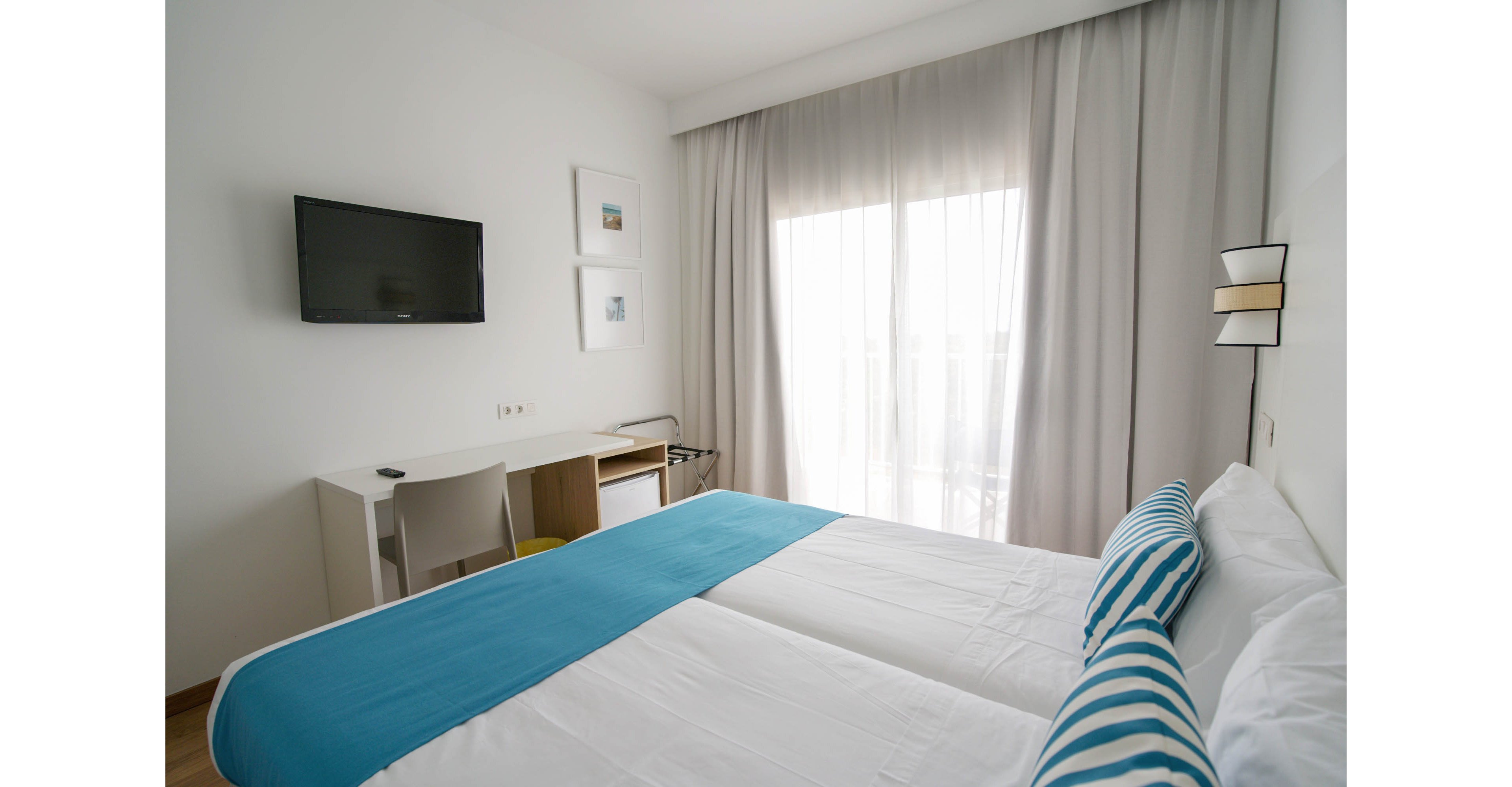 A clean, modern hotel room features a neatly made double bed with white linens and blue striped pillows. Sheer curtains cover a large window, allowing soft natural light to fill the space. A wall-mounted flat-screen television faces the bed above a simple white desk and chair. Minimalist decor and neutral walls create a calm, uncluttered atmosphere suitable for rest or work.