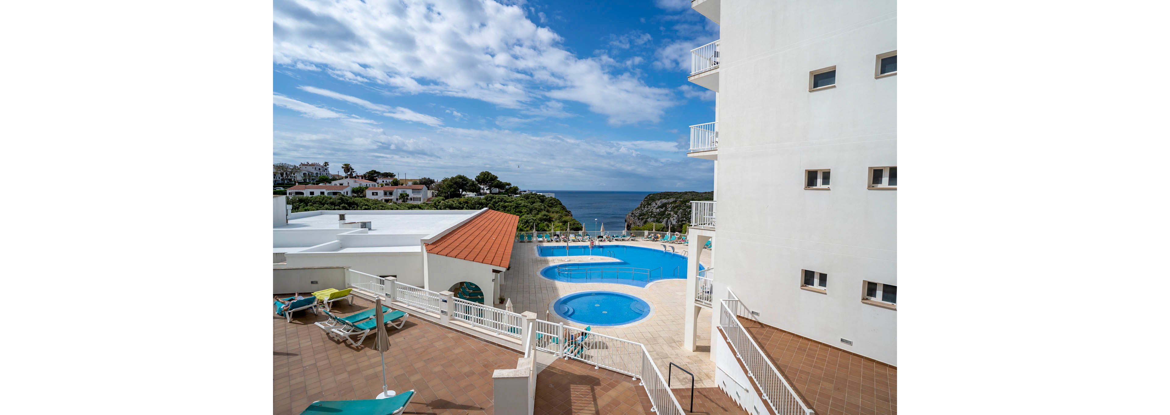 A seaside hotel complex features a spacious terrace with multiple circular swimming pools. Sun loungers and umbrellas are arranged around the pool area, suggesting a relaxing vacation setting. White modern buildings frame the view, leading the eye toward the blue ocean and partly cloudy sky in the distance. The scene conveys a bright, tranquil coastal resort atmosphere.