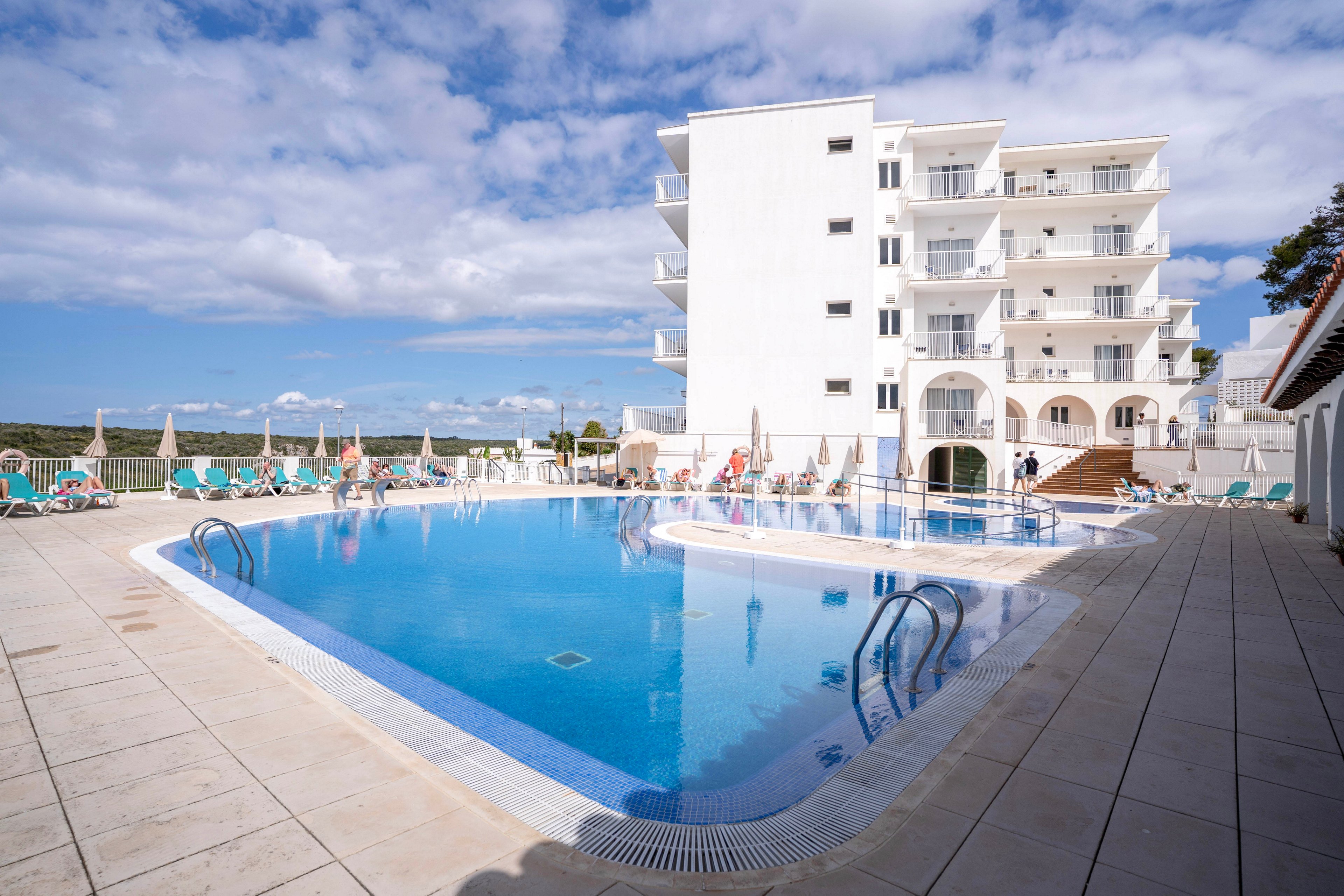 A large outdoor swimming pool sits in front of a multi-story white hotel building under a bright blue sky. Sun loungers and umbrellas line the pool deck, with a few people relaxing around the water. Stainless steel ladders lead into the clear pool, which reflects the surrounding architecture and clouds. The scene suggests a warm, leisurely vacation atmosphere at a coastal or hillside resort.