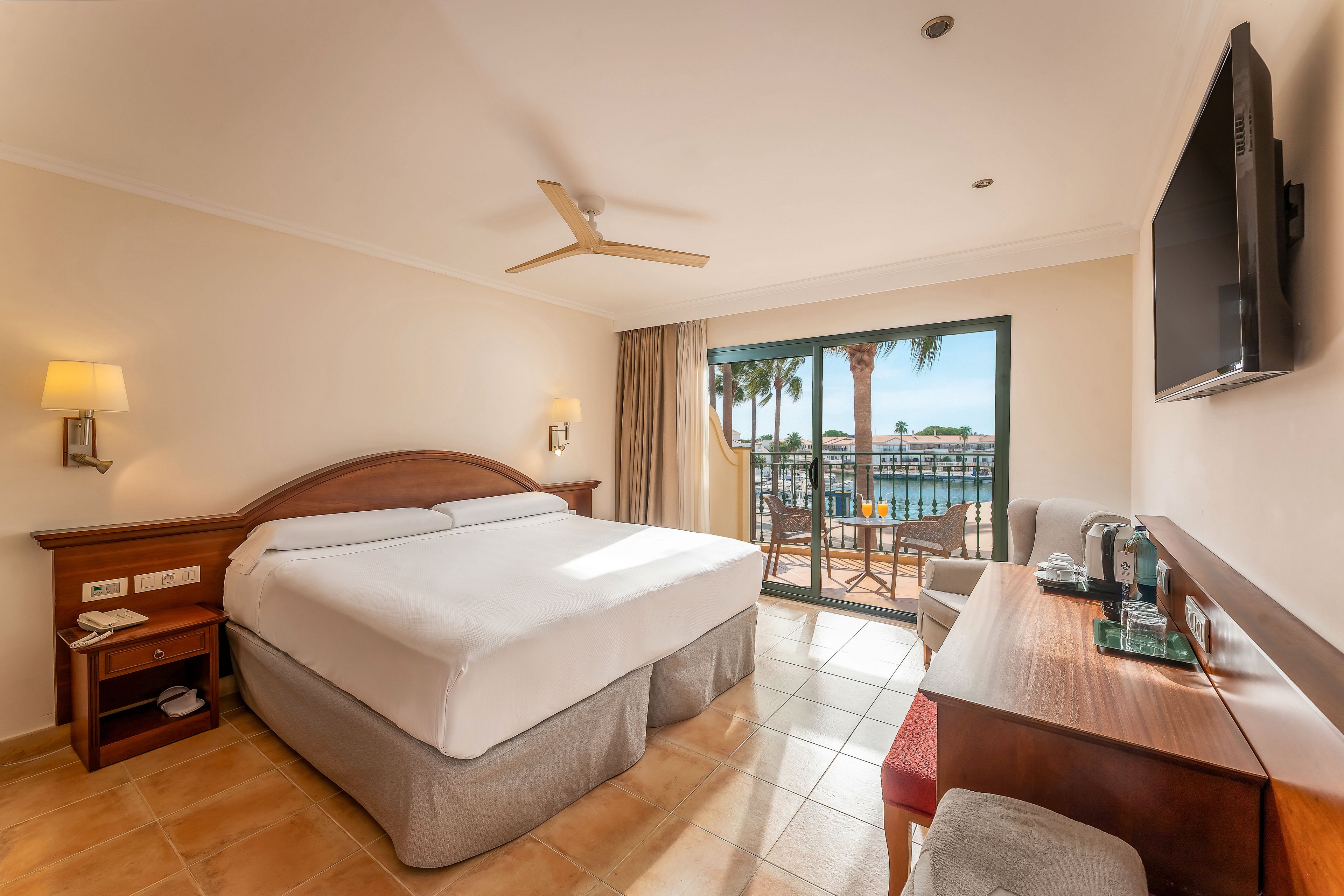 A neatly made double bed sits in a bright hotel room with tiled floors and neutral walls. A large glass door opens to a balcony overlooking palm trees and water. The room includes a wall-mounted television, wooden desk with chair, and bedside tables with lamps. Natural light fills the space, creating a warm and inviting atmosphere for guests.
