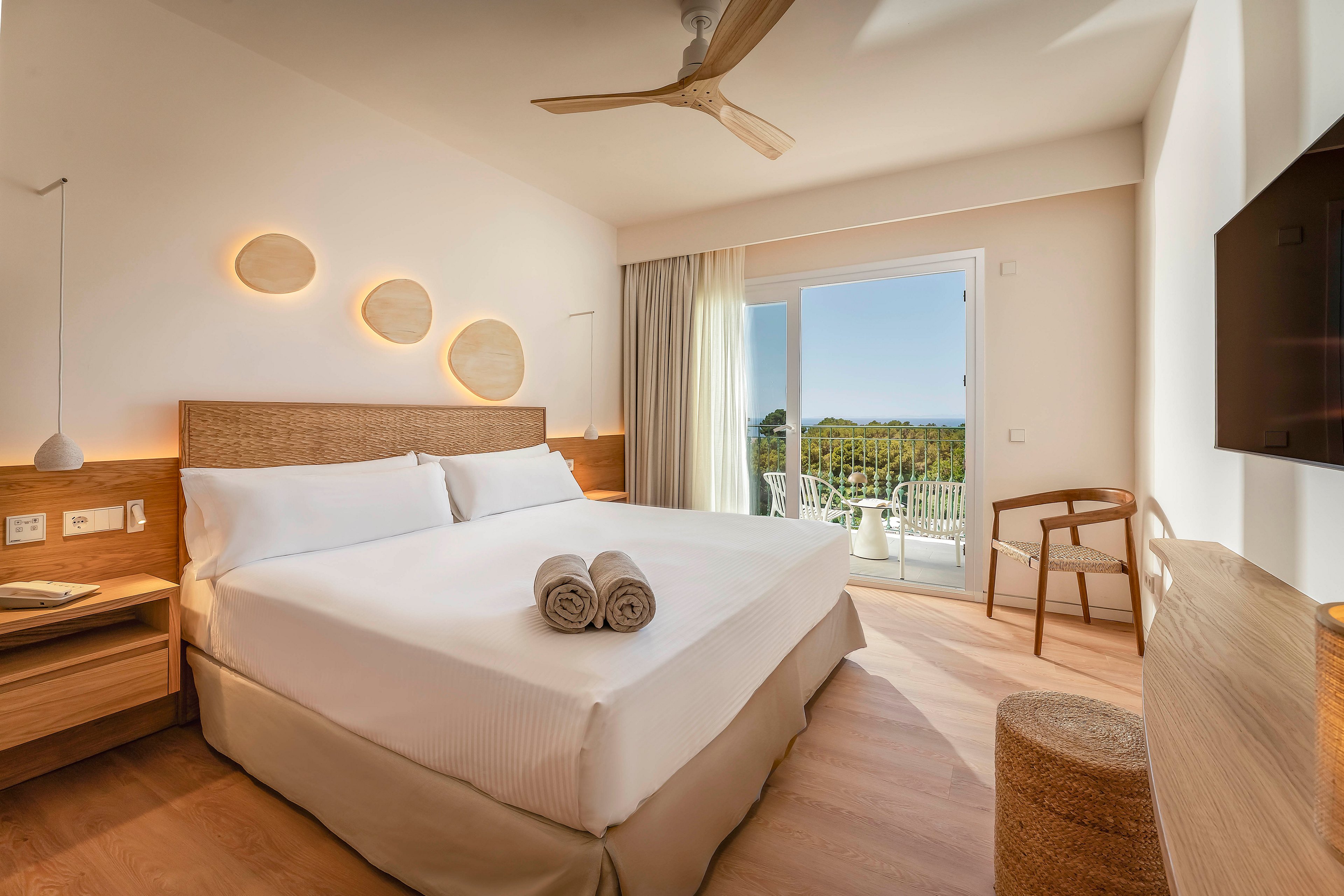 A contemporary hotel bedroom features a large neatly made bed with two rolled towels placed at the center. Warm wooden furniture, including bedside tables, a chair, and a woven stool, complement the light-toned flooring. Circular wall lights and a ceiling fan add to the minimalist decor, while sheer curtains frame a glass door leading to a balcony. Through the open door, a scenic landscape with greenery and blue sky is visible, enhancing the airy, relaxing atmosphere.