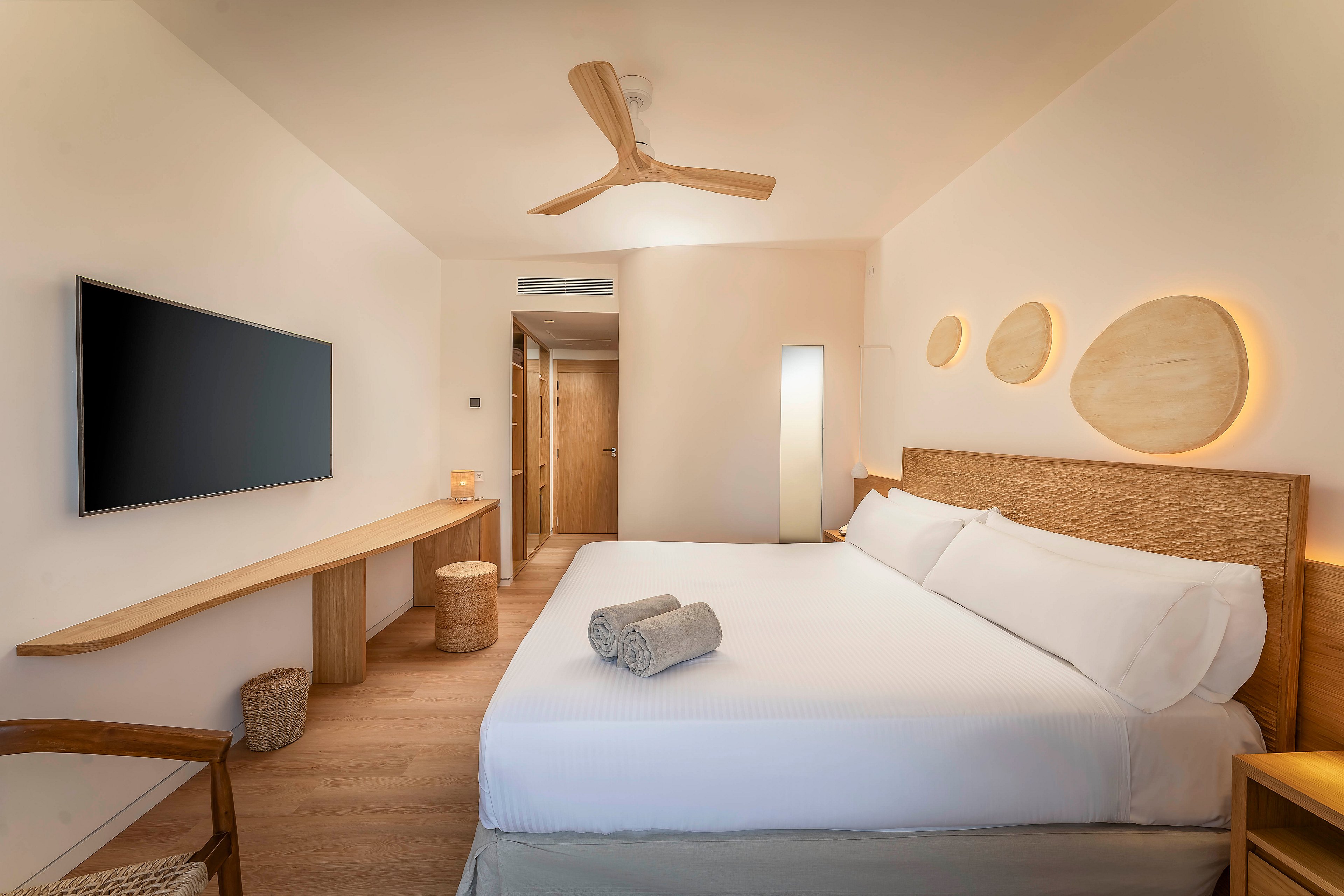 A contemporary hotel bedroom features a large neatly made bed with white linens and two rolled towels placed on top. The room is decorated in a minimalist style with light wood furniture, including a long desk, chair, and small stools. A flat-screen television is mounted on the wall opposite the bed, and circular wall accents with warm backlighting add visual interest. Soft neutral colors and indirect lighting create a calm, inviting atmosphere in the space.