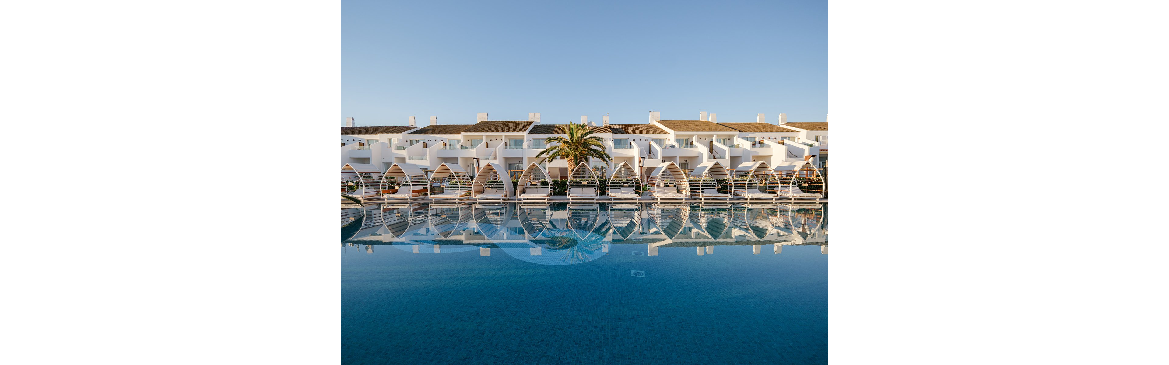A contemporary white resort complex stands behind a large, calm swimming pool that perfectly reflects its arched architecture. The scene is captured under clear blue skies, suggesting a warm, sunny day at a coastal destination. A single palm tree adds a tropical touch to the symmetrical composition. The overall atmosphere is tranquil and inviting, ideal for vacation or leisure imagery.