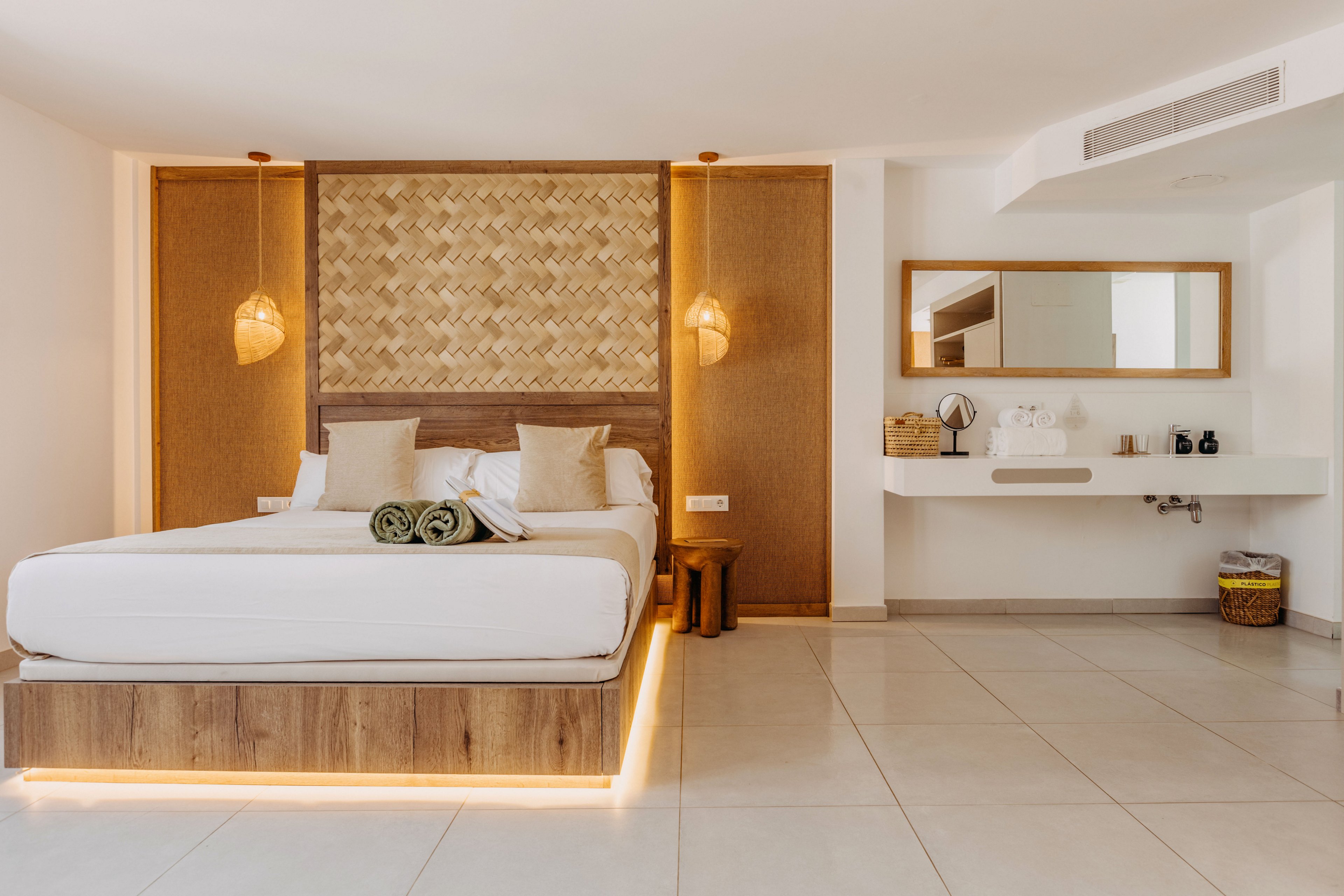 A contemporary hotel bedroom features a large bed with white linens and neutral-toned pillows. Warm wooden panels and woven textures frame the headboard, complemented by two hanging pendant lights. A floating vanity with a large mirror and decorative items sits along the right wall, adding to the clean, minimalist design. Soft under-bed lighting and spacious tiled flooring create an inviting, airy atmosphere.