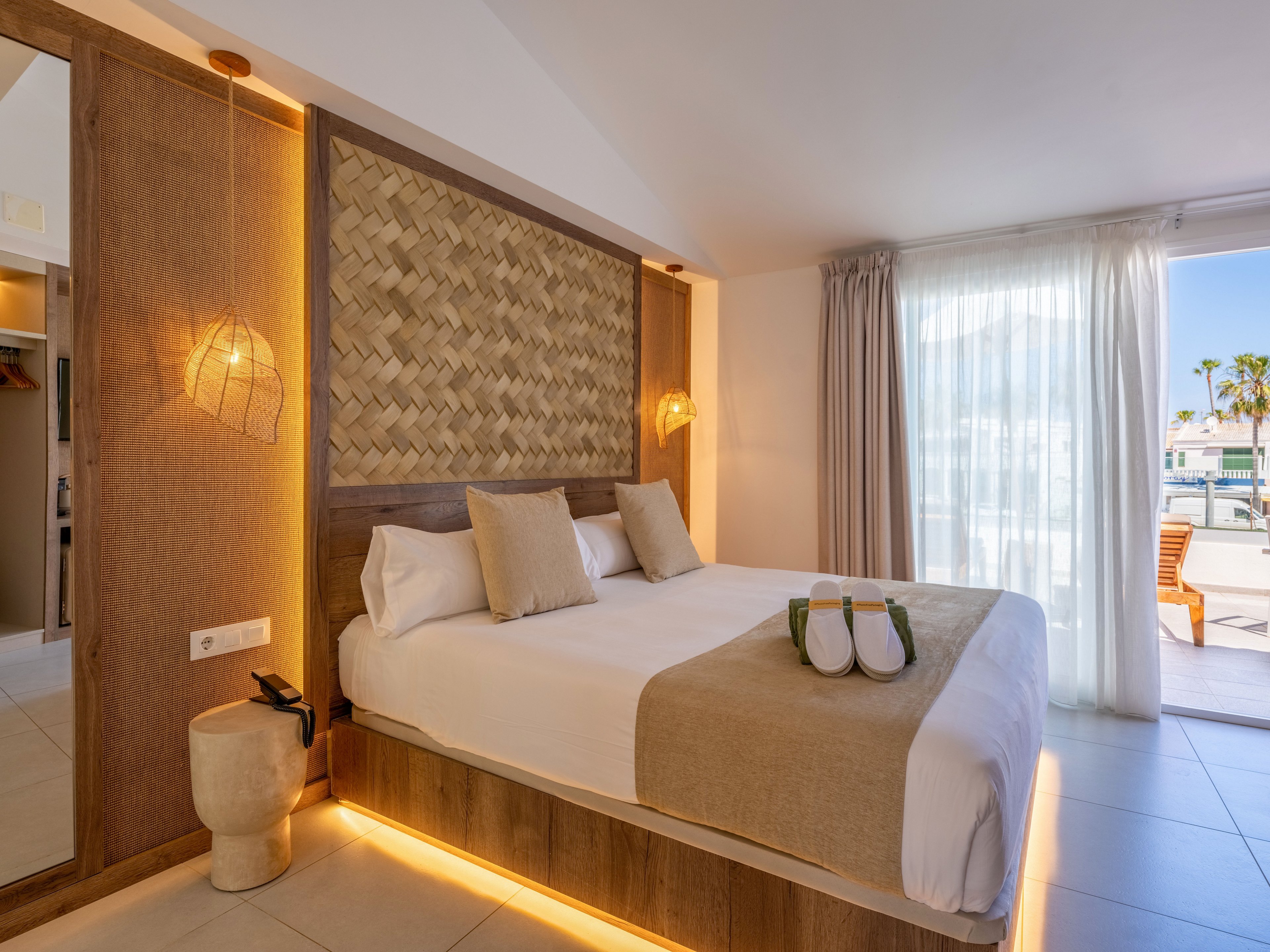 A contemporary hotel bedroom features a large bed with neutral linens and accent pillows. Warm wooden paneling, woven textures, and pendant lights create a cozy, minimalist atmosphere. Soft under-bed lighting and natural daylight from the large window enhance the airy feel of the space. Outside, a terrace with seating and palm trees suggests a coastal or resort setting.