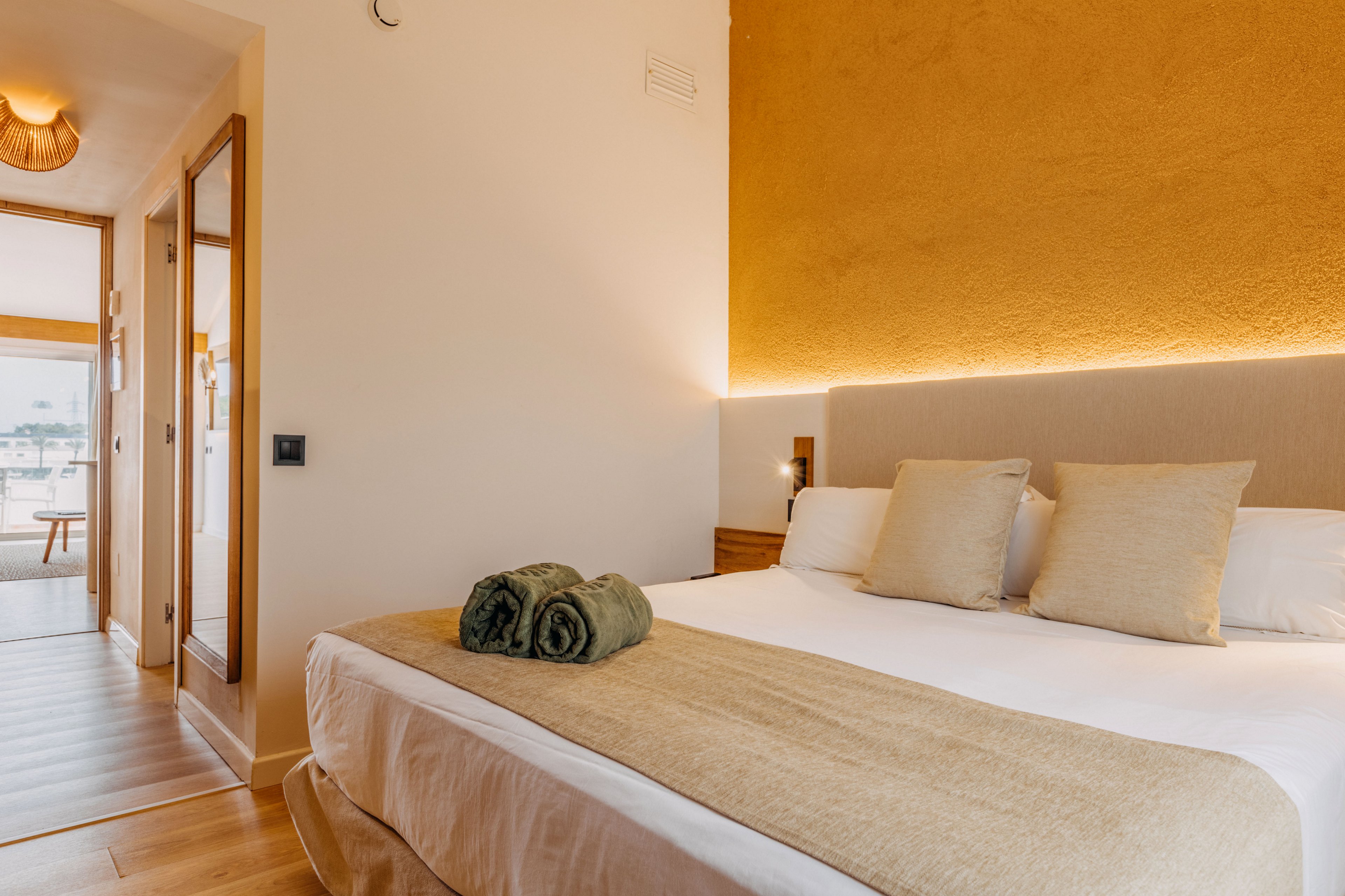 A neatly made double bed with neutral-toned linens and pillows is the focal point of this modern hotel room. Rolled green towels rest at the foot of the bed, adding a subtle accent of color. Warm ambient lighting highlights a textured mustard-colored wall behind the headboard. A hallway with wooden flooring leads to a bright sitting area and balcony, suggesting a comfortable and inviting stay.