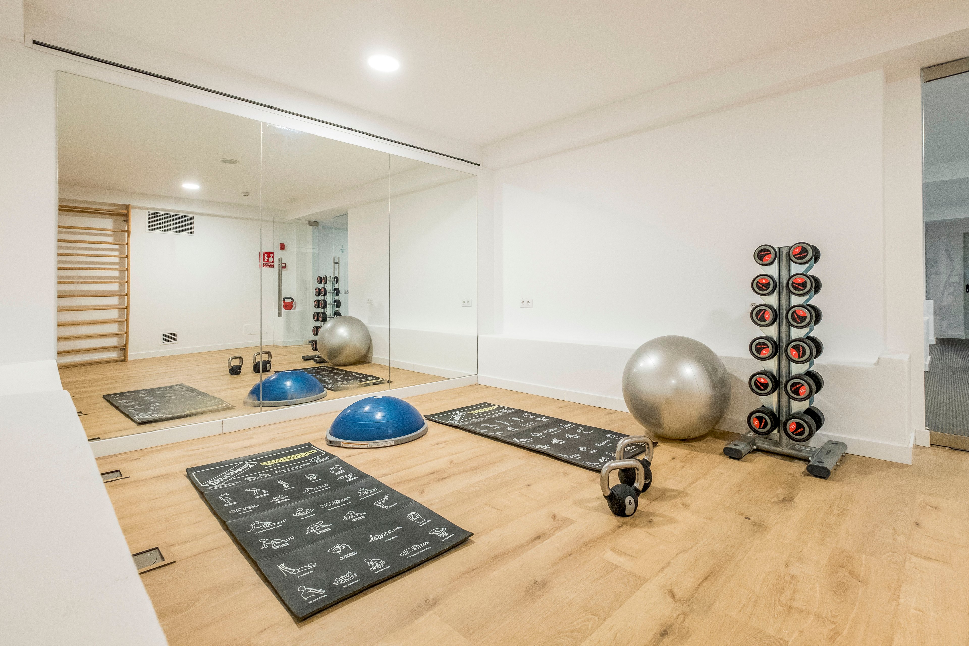Bright, minimalist fitness room featuring various exercise equipment on a light wooden floor. Two black workout mats, a blue balance trainer, kettlebells, and a large silver stability ball are arranged in front of a wall mirror. A vertical rack of dumbbells stands to the right, while wooden wall bars and a fire extinguisher are visible in the background. The clean white walls and ample lighting create a fresh, contemporary workout environment.