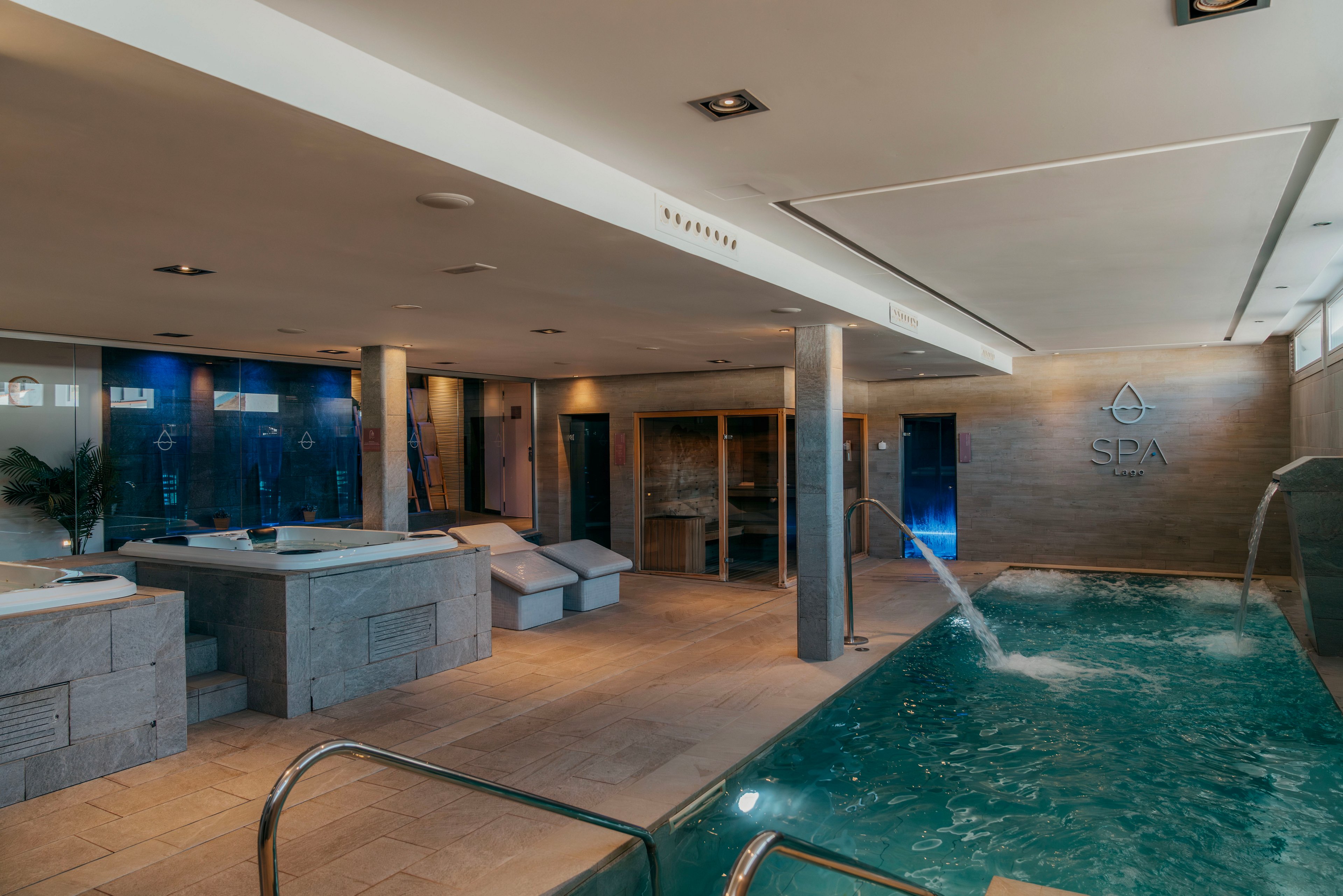 A contemporary indoor spa features a turquoise hydrotherapy pool with water jets and stainless steel handrails. Stone-tiled floors and walls create a warm, natural atmosphere throughout the space. Several treatment beds and lounge chairs are arranged around the pool for relaxation. Soft, indirect lighting and clean architectural lines emphasize a calm, luxurious wellness environment.