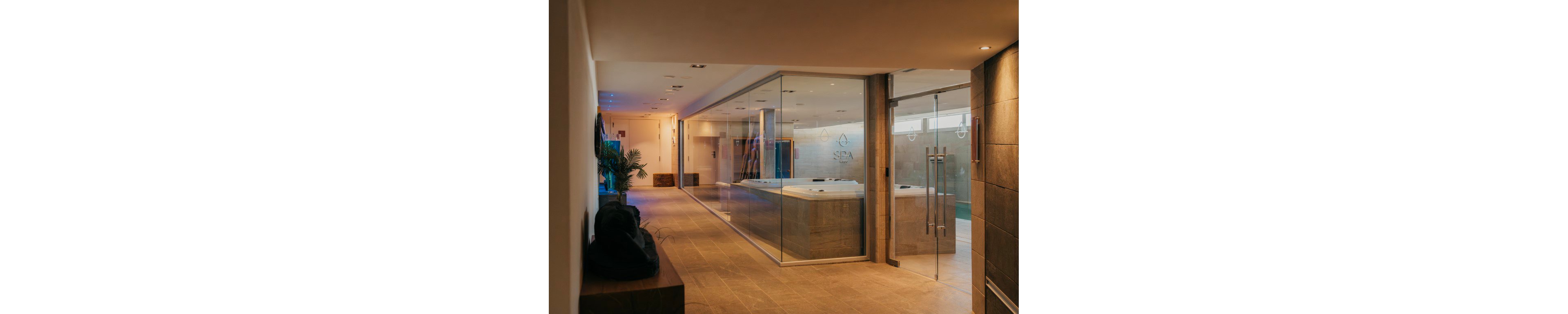 A contemporary spa interior features a large glass-enclosed jacuzzi area along a warm, tiled corridor. Soft ambient lighting and clean architectural lines create a calm, luxurious atmosphere. The space includes glass doors, minimalist decor, and subtle wall graphics with water-themed icons. Potted plants and neutral tones enhance the relaxing wellness setting.