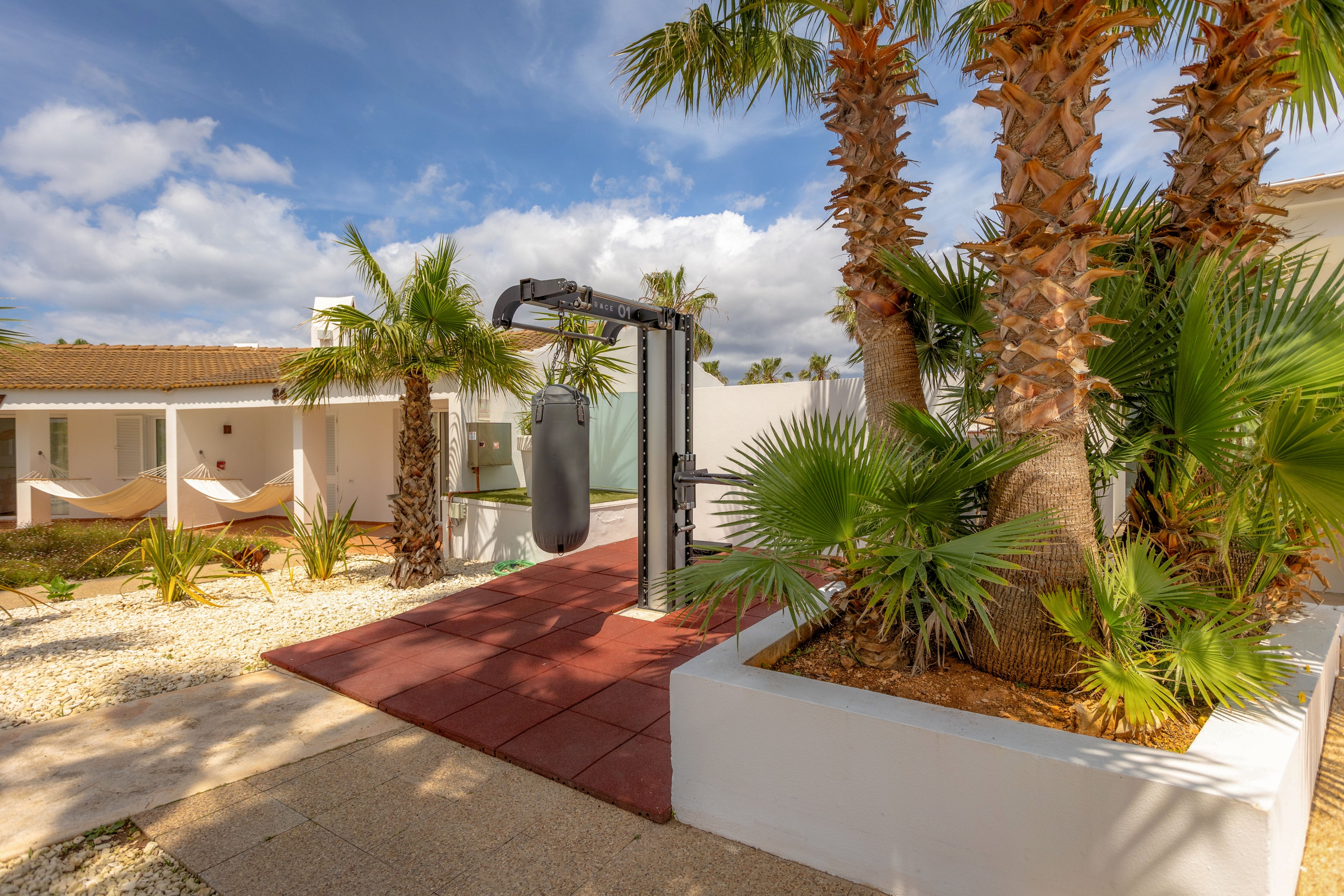 An outdoor punching bag stands on red rubber tiles in a modern resort courtyard. The scene is surrounded by palm trees, white walls, and gravel landscaping under a partly cloudy sky. A hammock is visible near the building, suggesting a mix of relaxation and fitness. The overall atmosphere is bright, warm, and inviting for vacation exercise.