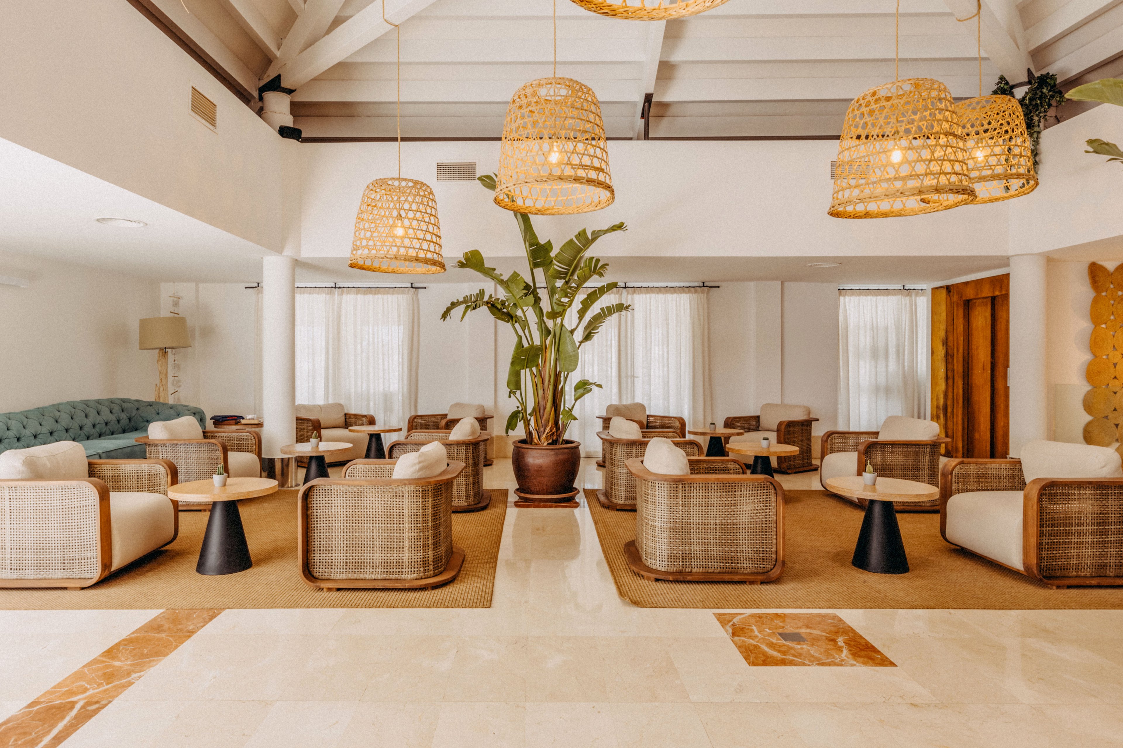 Spacious lounge area featuring multiple wicker armchairs arranged around small black side tables. The setting appears to be a contemporary hotel or resort lobby with large windows and sheer curtains letting in soft natural light. Warm woven pendant lights hang from the ceiling, complementing the neutral tones of the tiled floor and walls. A large potted plant adds greenery, enhancing the relaxed, inviting atmosphere.
