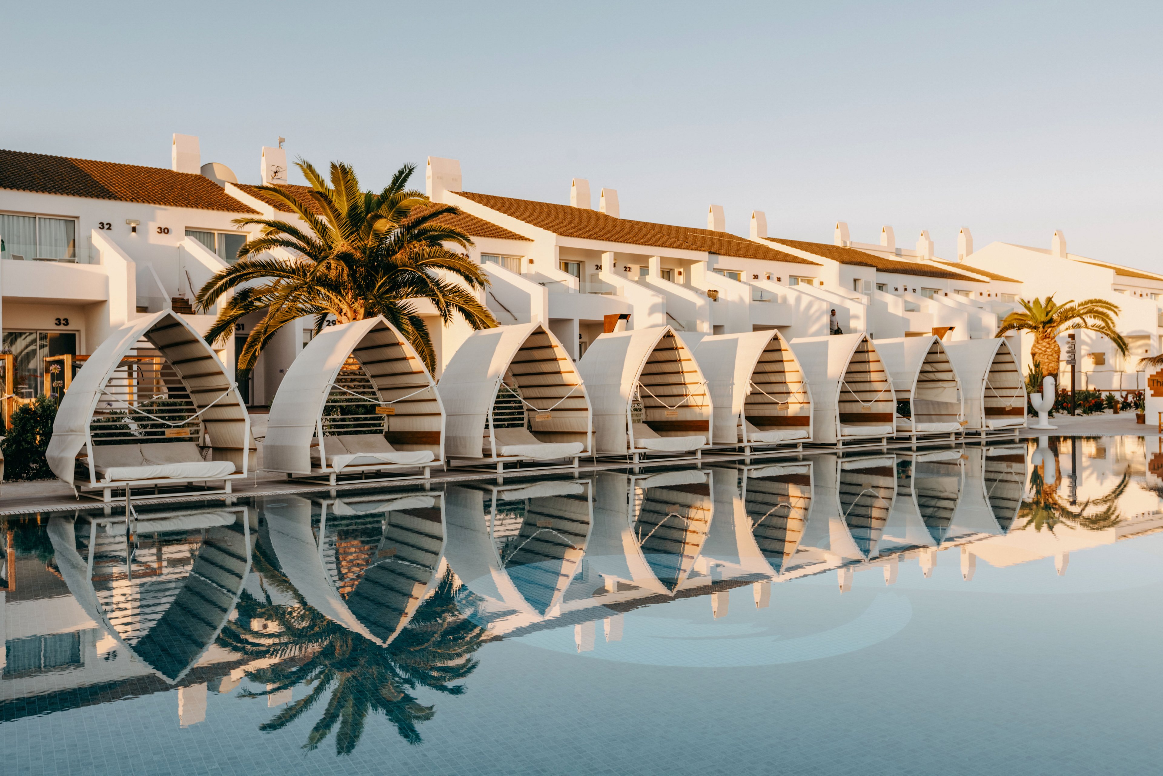 A row of contemporary white cabanas lines the edge of a calm swimming pool at a modern seaside resort. The architecture features curved, sail-like structures in front of multi-level white buildings. Palm trees add a tropical touch, and the clear water reflects the cabanas and warm evening light. The overall scene suggests a tranquil vacation destination with stylish outdoor lounge areas.