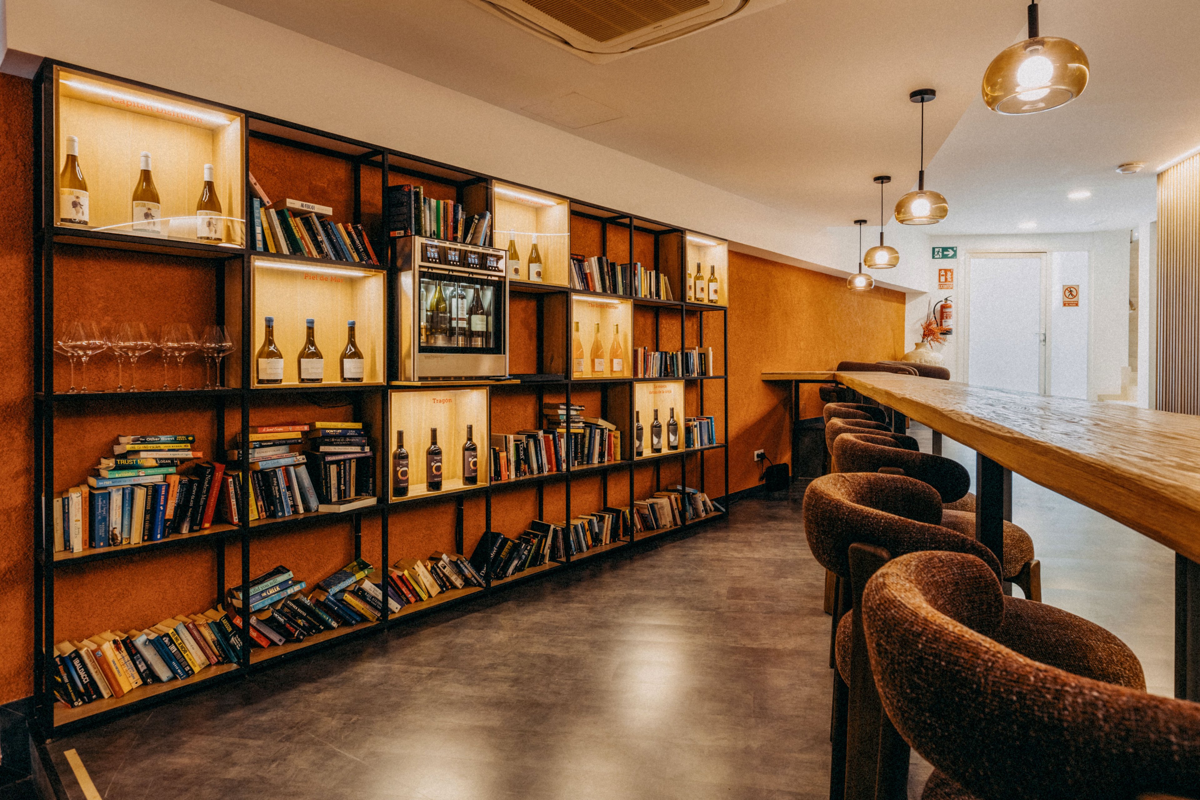 A stylish bar lounge features a long wooden counter with upholstered stools and warm pendant lighting. Along one wall, an illuminated shelving unit displays wine bottles, glassware, and rows of colorful books. The polished concrete floor and minimalist ceiling fixtures create a modern yet inviting atmosphere. The scene suggests a relaxed social space that blends a library aesthetic with a contemporary bar setting.