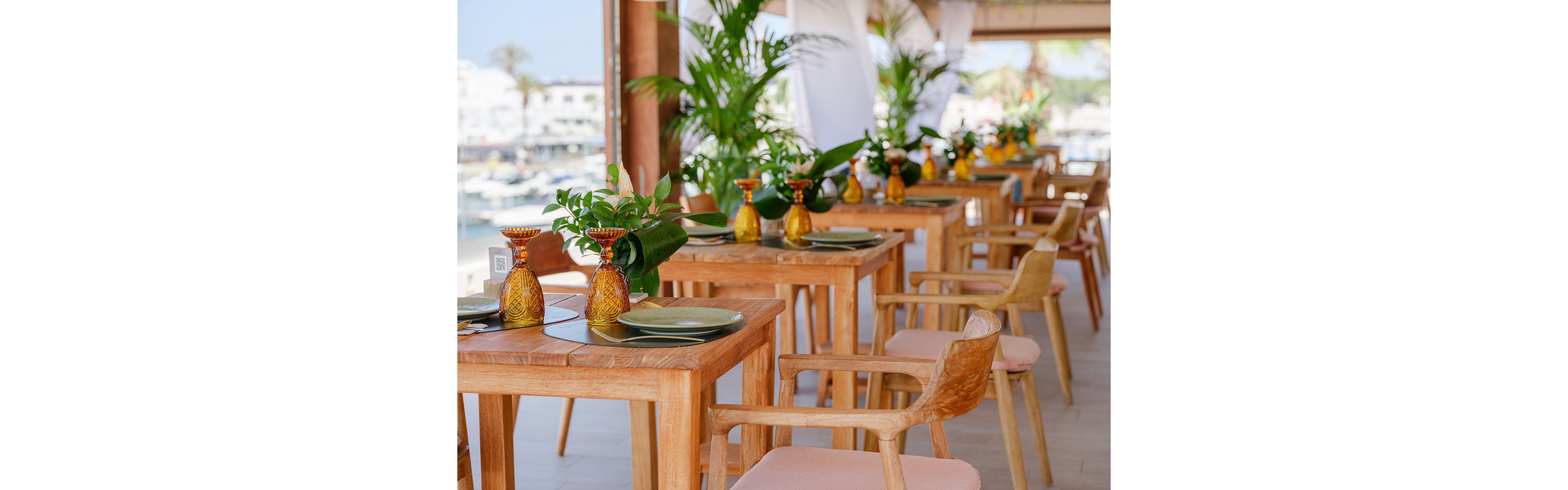 A bright open-air restaurant terrace features rows of wooden tables and chairs neatly arranged for guests. Each table is set with green plates, decorative amber glass vases, and lush green foliage centerpieces. Large potted plants and white fabric drapes enhance the tropical atmosphere, while a marina and waterfront buildings are visible in the background. The overall scene feels inviting, relaxed, and ideal for outdoor dining by the sea.