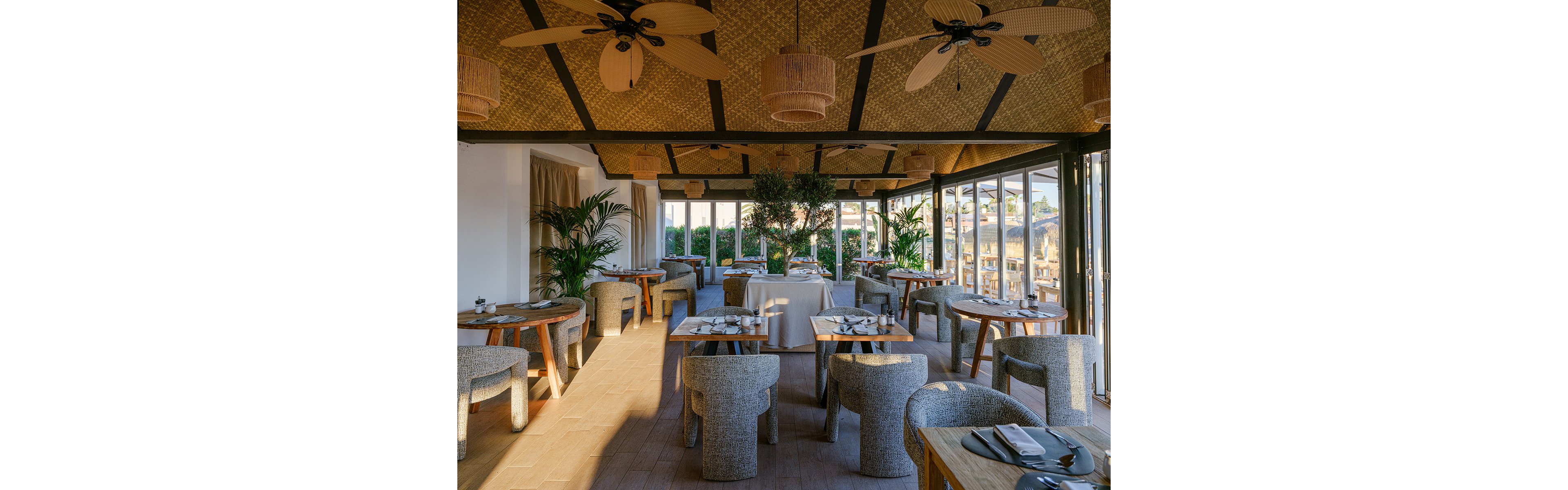 Contemporary restaurant dining room with round wooden tables and textured upholstered chairs is bathed in warm natural light. Large floor-to-ceiling glass doors open to an outdoor terrace, offering views of surrounding buildings and greenery. Ceiling fans and woven light fixtures complement the wood and rattan finishes, creating a relaxed, coastal atmosphere. Potted plants and neatly set tables with plates and cutlery add to the inviting, upscale ambiance.