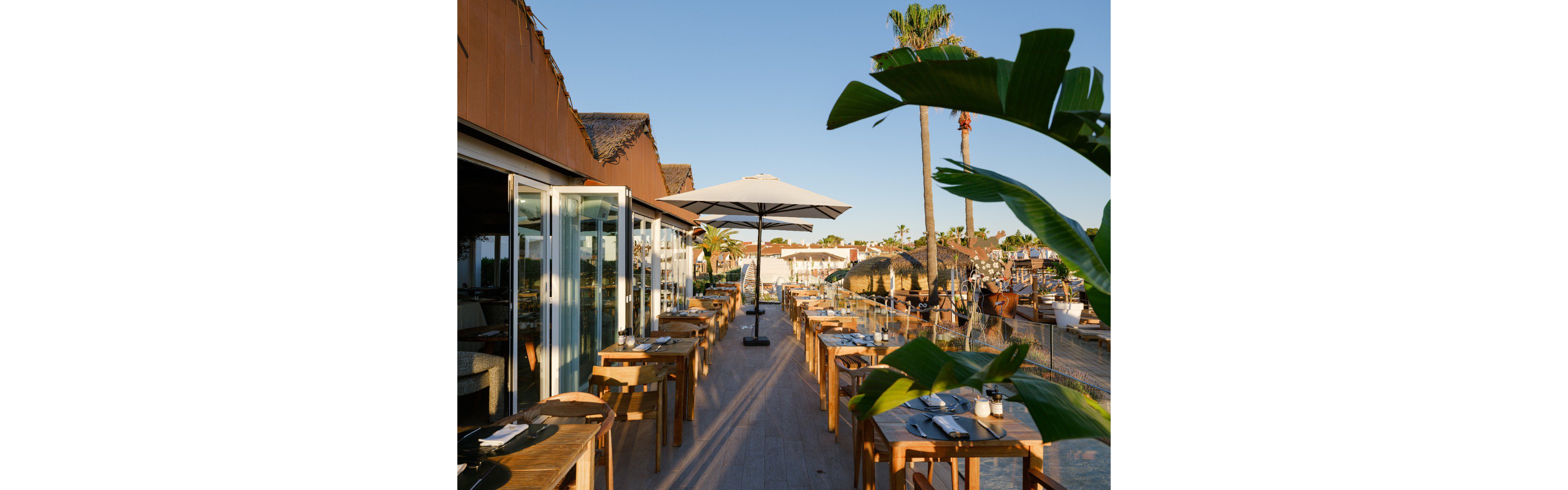 An open-air restaurant terrace is set with wooden tables and chairs, each prepared with plates, cutlery, and glassware. A large white umbrella stands centrally along the walkway, while glass doors connect the terrace to the interior dining area. Palm trees, thatched roofs, and distant buildings suggest a coastal or resort setting under warm evening light. The scene feels relaxed and inviting, ideal for al fresco dining by the water.