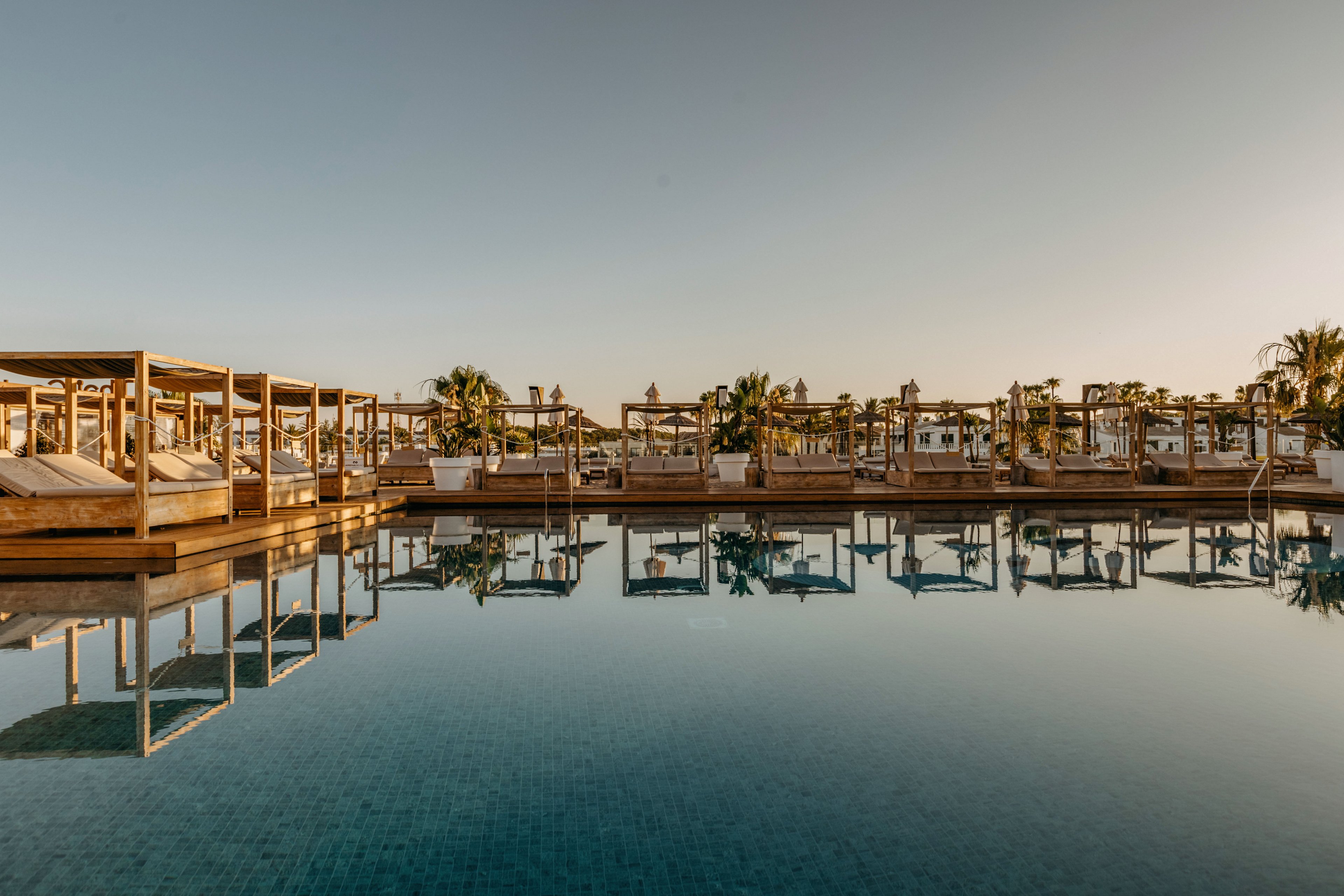 A tranquil resort swimming pool reflects rows of wooden cabanas and palm trees in the calm water. The scene is captured at sunset or early evening, with warm golden light and a clear sky. Cushioned lounge beds under pergola-style cabanas suggest a relaxing, upscale vacation atmosphere. The overall mood is serene and inviting, emphasizing leisure and waterfront relaxation.