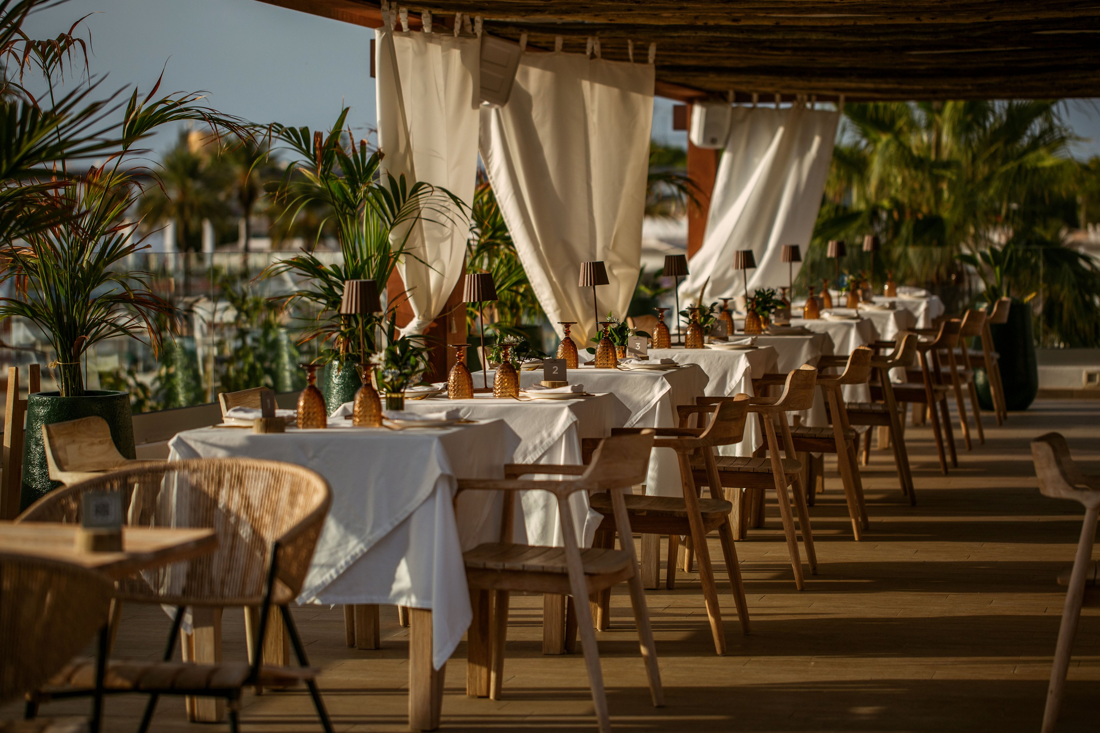 An elegant outdoor restaurant terrace is set with rows of wooden tables covered in white tablecloths. Natural wicker chairs and warm-toned table lamps create a cozy, inviting atmosphere. Tall green plants and flowing white curtains frame the space, suggesting a seaside or resort setting. Soft golden light enhances the relaxed, upscale dining mood.