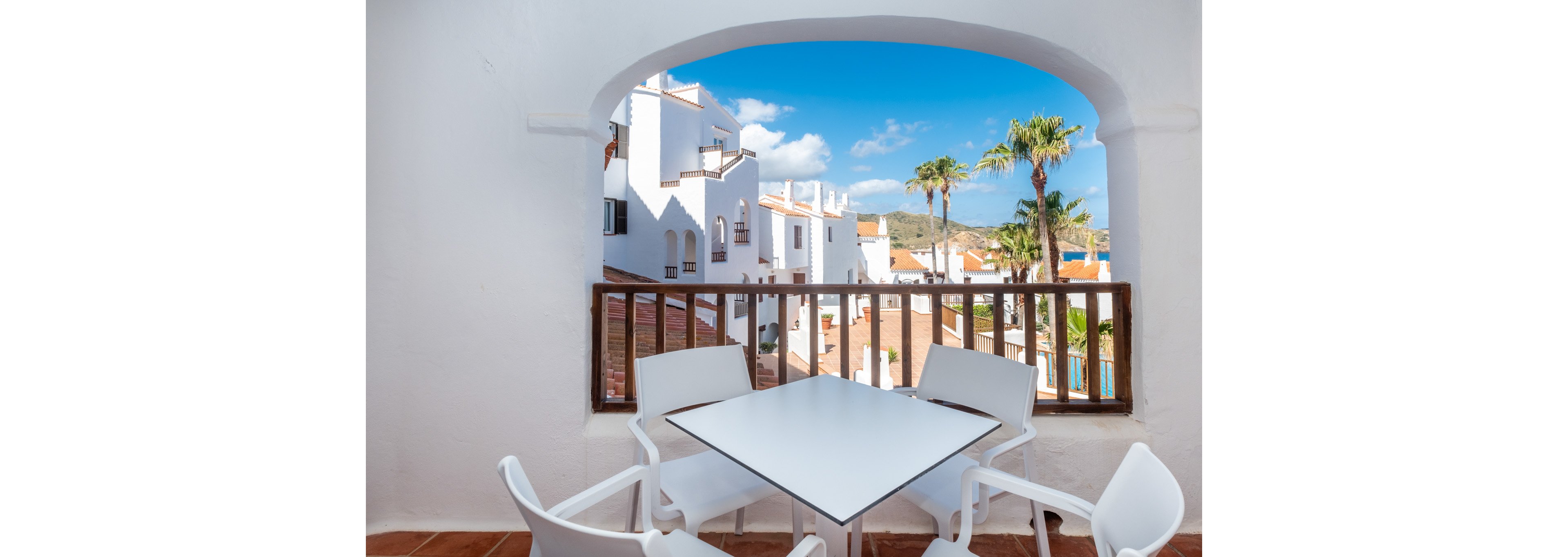 A bright terrace with a modern square table and four white chairs opens onto a view of whitewashed buildings. The scene is framed by an arched wall and wooden railing, creating a cozy outdoor seating area. Palm trees, tiled roofs, and distant hills suggest a warm Mediterranean or coastal town. Clear blue sky and vivid sunlight give the image a relaxed vacation atmosphere.