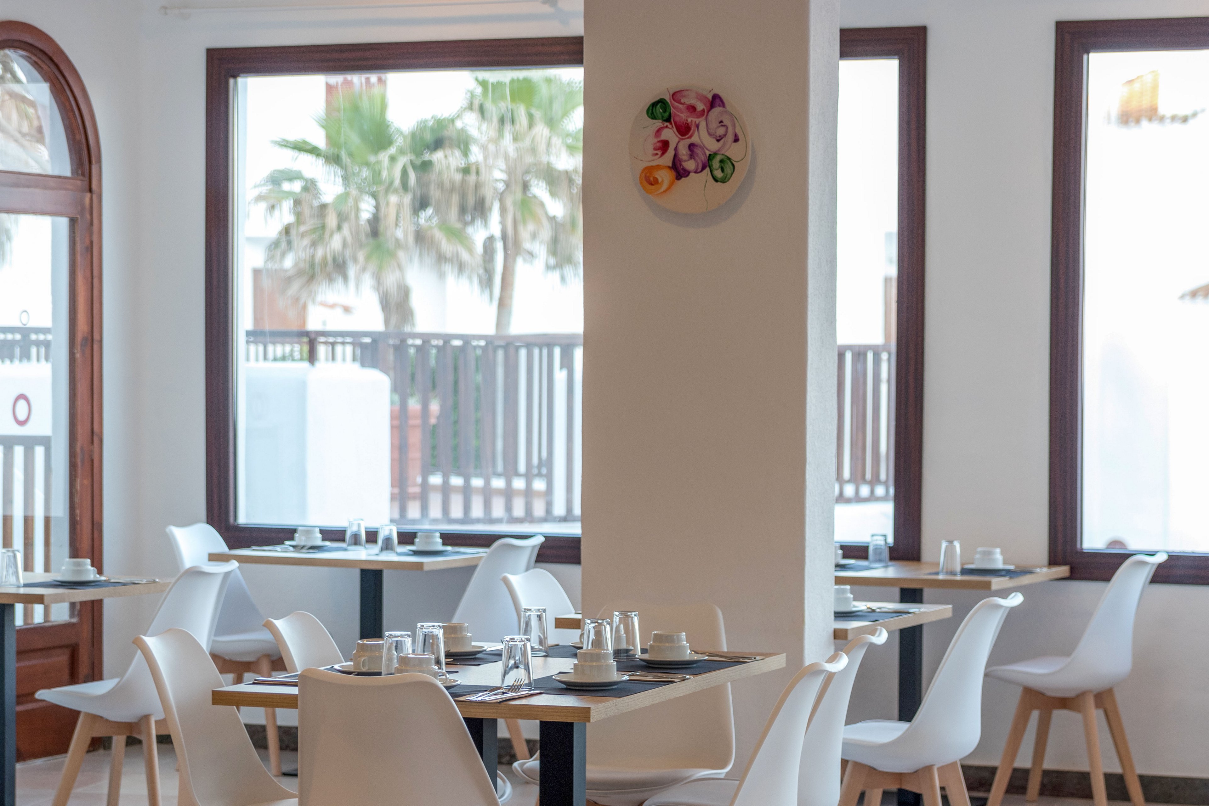 A contemporary restaurant dining area features minimalist white chairs and light wooden tables neatly set with glasses and cutlery. Large windows let in abundant natural light and reveal an outdoor view with palm trees and a wooden fence. A central white column divides the space and displays a colorful circular wall art piece. The overall atmosphere is clean, airy, and relaxed, suitable for casual dining or a quiet meal.