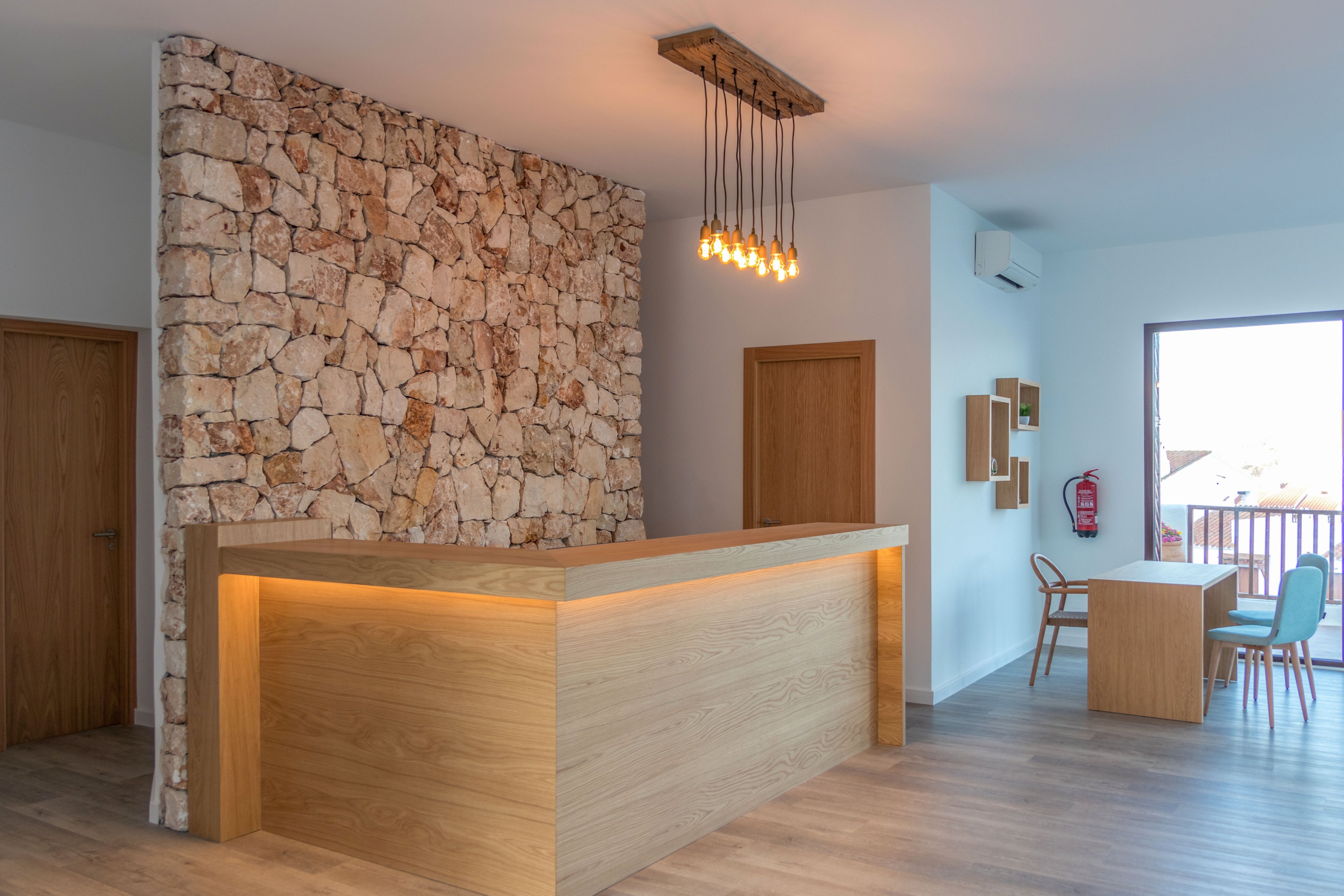 Contemporary interior space features a minimalist wooden reception desk with warm underlighting. A textured stone accent wall and wooden doors frame the area, creating a natural, welcoming atmosphere. Pendant lights hang from the ceiling above the desk, while a small seating area with table and chairs is visible near a balcony. Clean lines, neutral tones, and ample natural light emphasize the modern design.