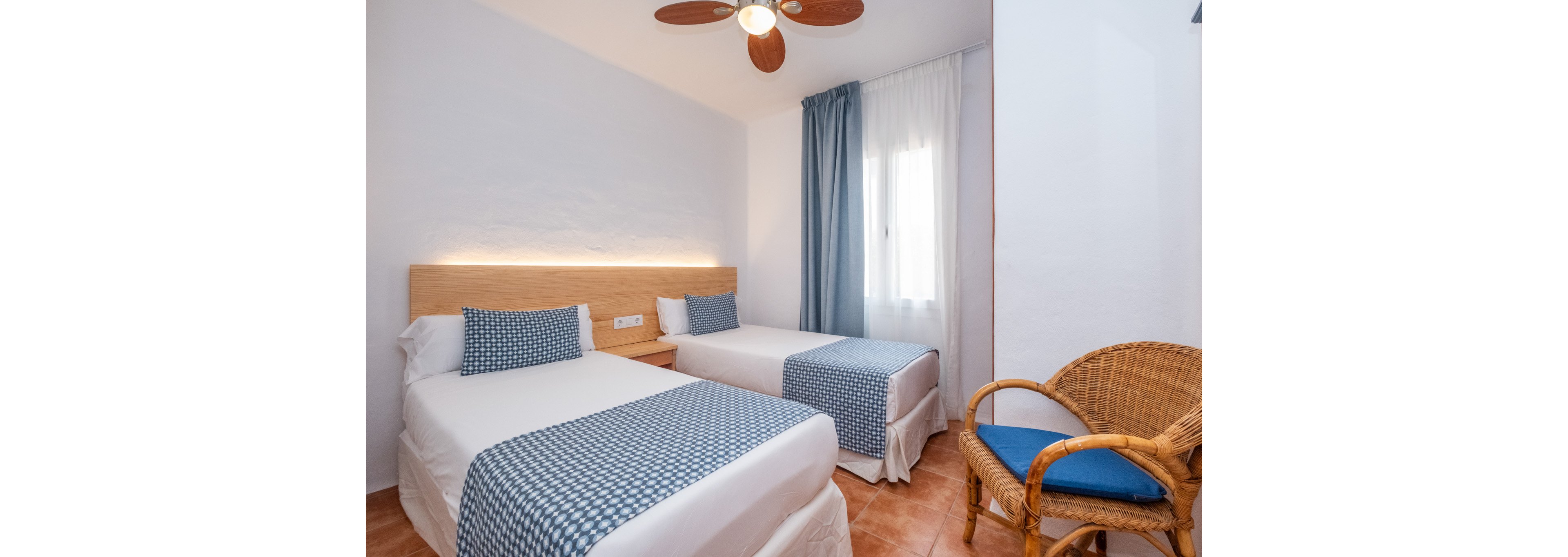 A cozy bedroom features two single beds with white linens and blue patterned throws and pillows. The room has a simple wooden headboard with soft backlighting and a tiled floor. A wicker chair with a blue cushion sits in the corner, and a window with sheer white and blue curtains lets in natural light. A ceiling fan with wooden blades hangs above, adding to the airy, relaxed atmosphere.