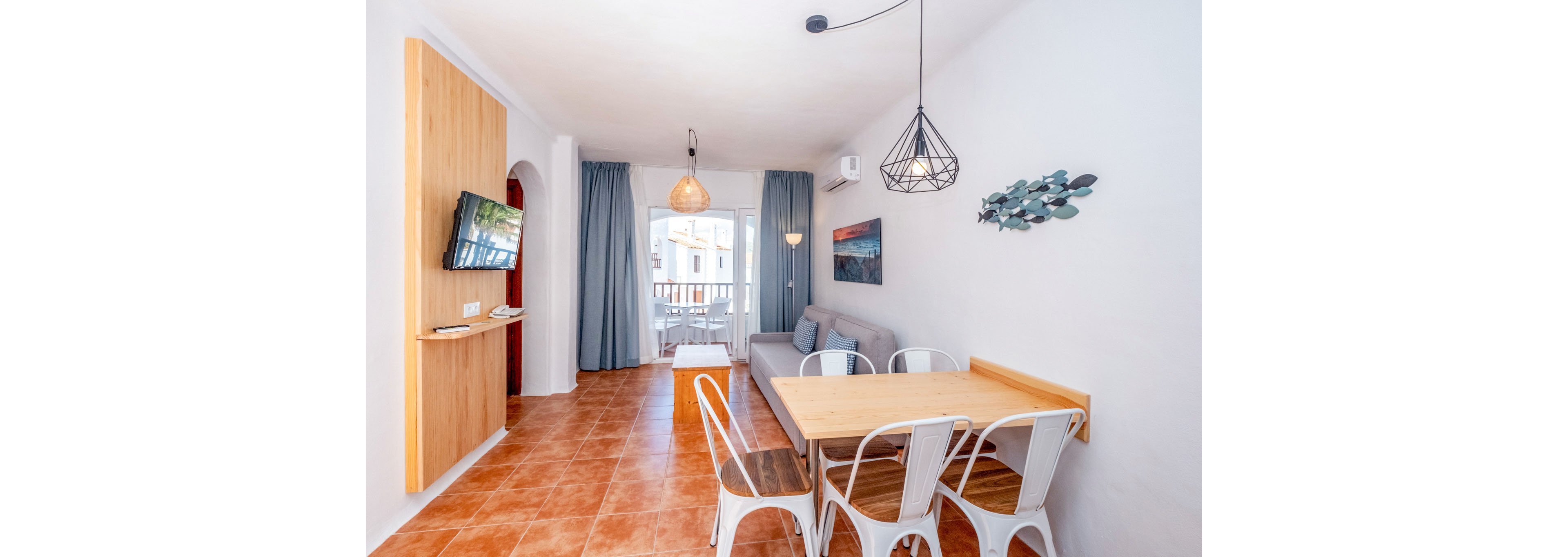 Sunlit open-plan apartment interior features a small dining table with white metal chairs and a cozy gray sofa. Terracotta floor tiles and white walls create a warm, clean atmosphere. Pendant lights hang from the ceiling, and a wall-mounted television faces the seating area. Large glass doors with blue curtains open to a balcony, revealing neighboring buildings and outdoor light.