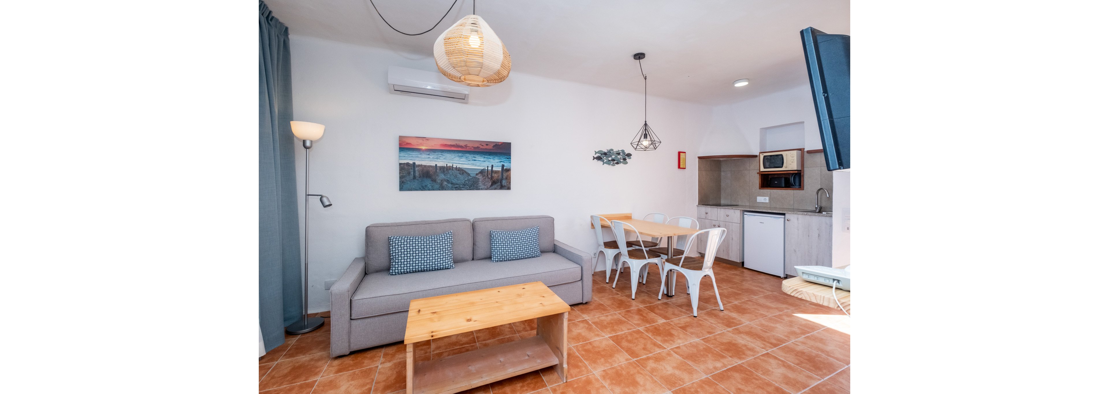 Cozy modern living room features a gray sofa with patterned cushions and a wooden coffee table on terracotta floor tiles. A small dining area with a wooden table and white metal chairs sits beside a compact kitchenette with microwave, sink, and mini fridge. Wall art, pendant lights, and a floor lamp add decorative touches to the white walls. The space appears clean, bright, and inviting, suitable for a vacation rental or small apartment.