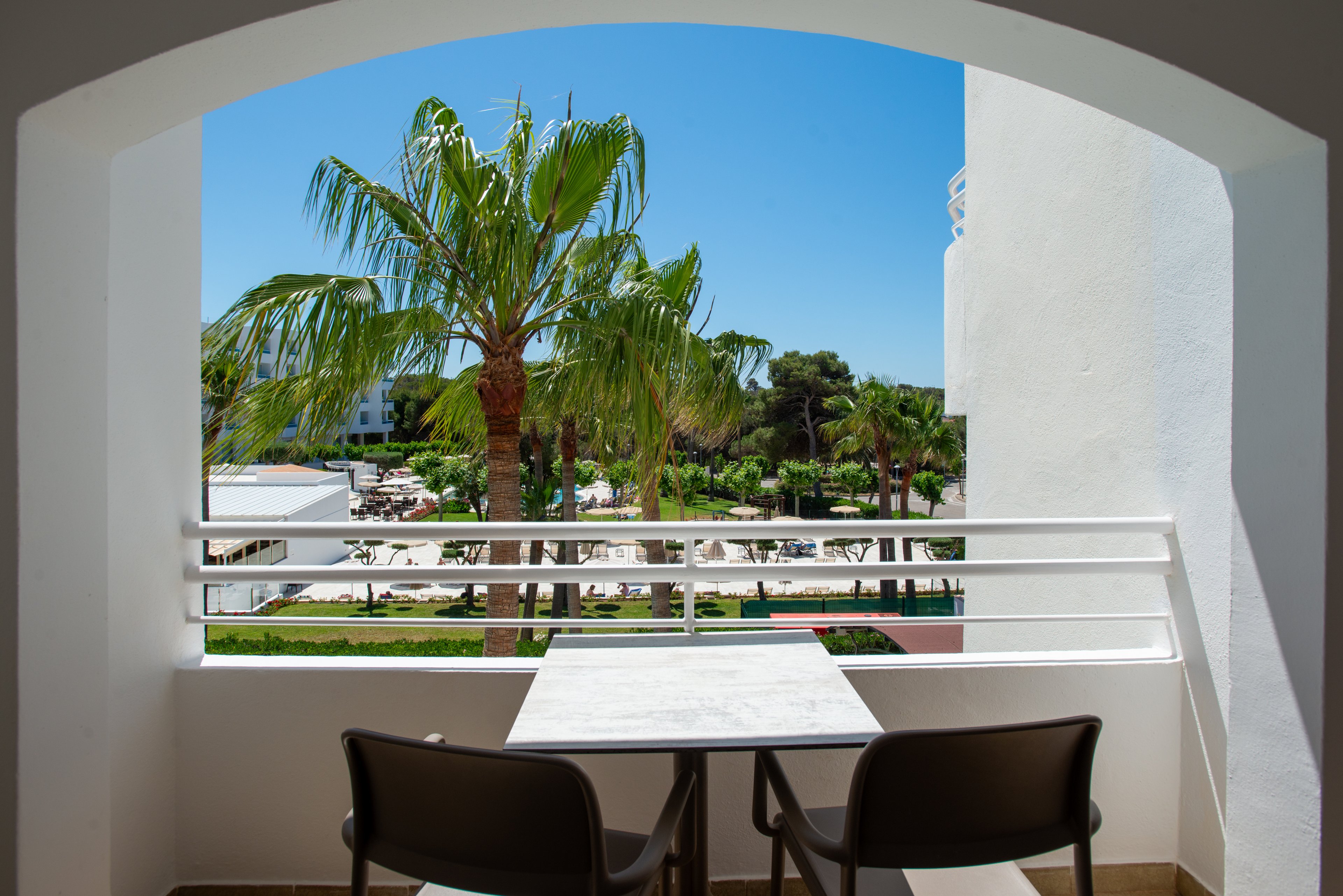 A small balcony with a white table and two dark chairs overlooks a lush resort garden. Tall palm trees rise above green lawns and pathways, with white buildings visible in the background. The scene is framed by a white archway and railing, creating a relaxed holiday atmosphere. Bright sunlight and a clear blue sky suggest a warm, sunny day in a coastal or tropical destination.