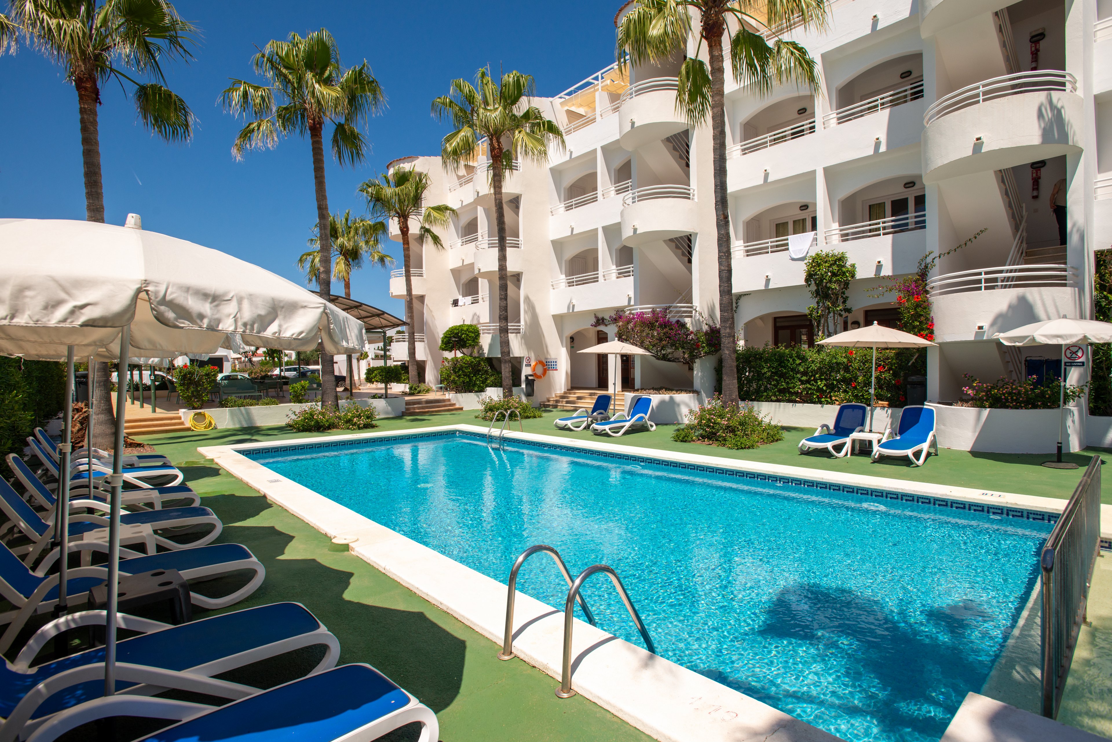 Outdoor swimming pool at a bright white resort is surrounded by blue lounge chairs and umbrellas. Tall palm trees frame the scene, adding a tropical atmosphere under a clear blue sky. The multi-story building features balconies overlooking the pool area, suggesting vacation accommodations. The overall setting feels relaxed and inviting, ideal for leisure and sunbathing.