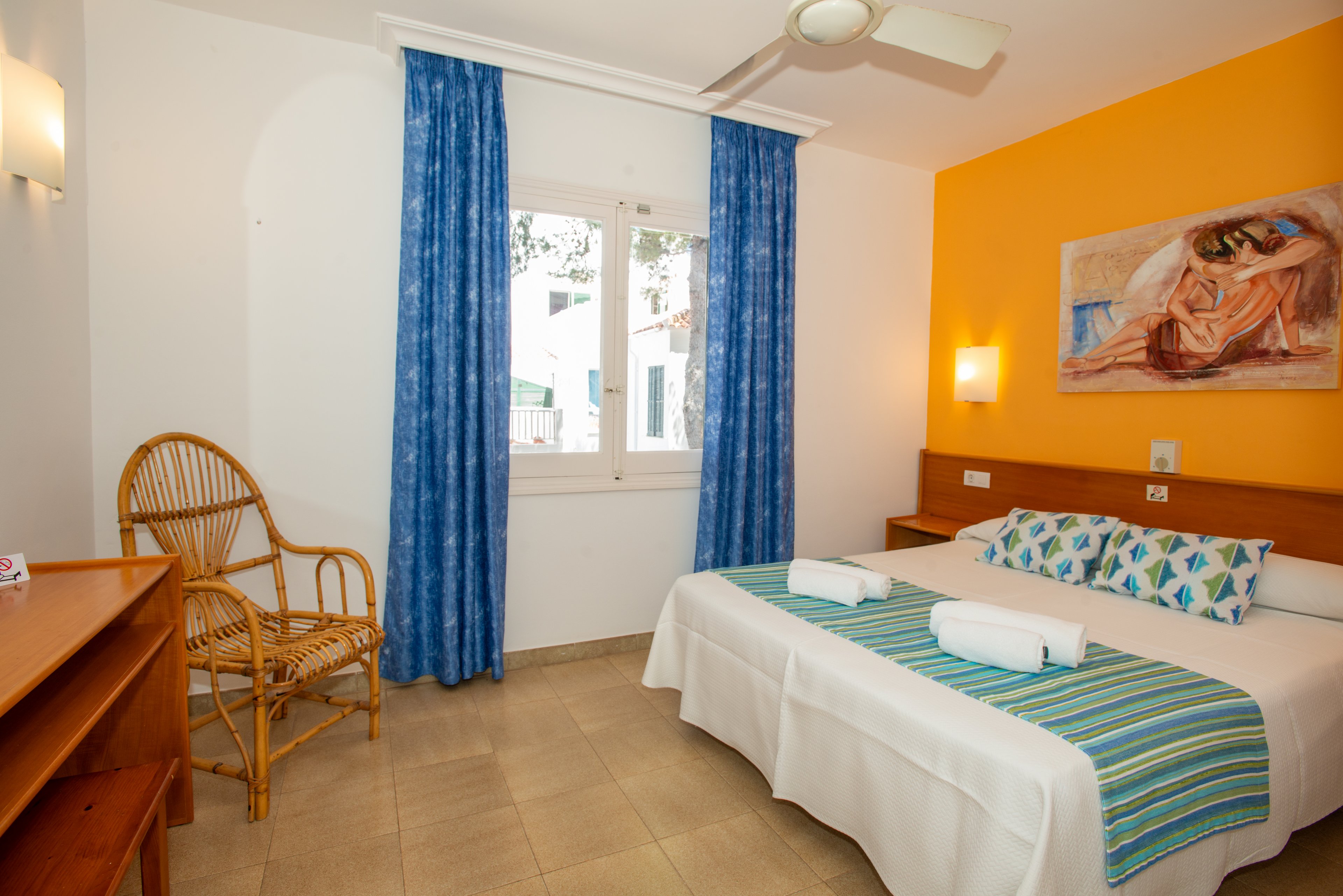 A tidy hotel bedroom features a double bed with white linens and patterned pillows against a vibrant yellow accent wall. Two rolled white towels rest on a striped blue and green bed runner. The room includes a wooden desk, wicker chair, and bedside tables with wall-mounted lights. Blue curtains frame a window that lets in natural light, and a ceiling fan hangs above for comfort.