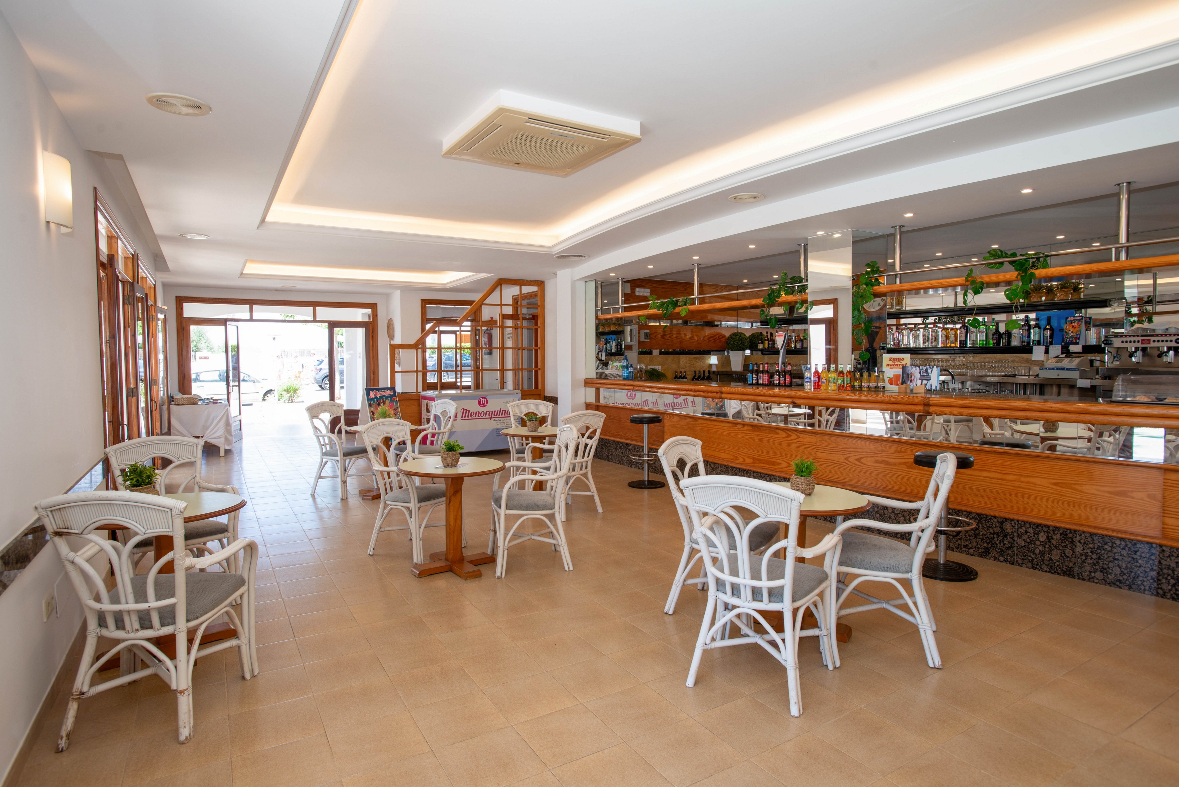 Spacious cafe interior featuring a long wooden bar counter and neatly arranged bottles and glassware. White wicker chairs surround small round tables with potted plants, creating a casual seating area. Large windows and glass doors let in abundant natural light, highlighting the clean tiled floor. The overall atmosphere is airy, contemporary, and welcoming, suitable for relaxed dining or drinks.